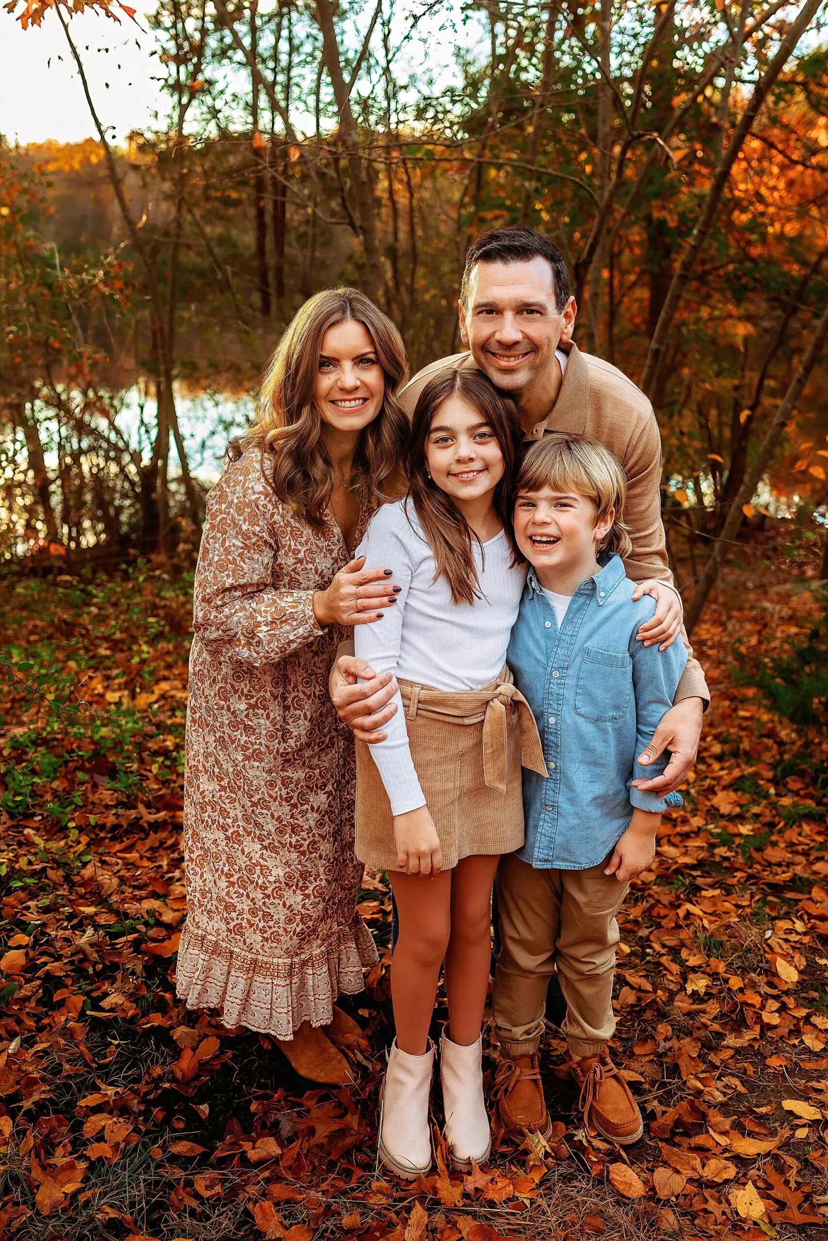 A smiling family of four in Connecticut stands outdoors on fallen autumn leaves. The woman wears a patterned dress, the man a beige sweater, the girl a white top and tan skirt, and the boy a blue shirt. Trees and a lake provide the perfect backdrop for their family photos session.