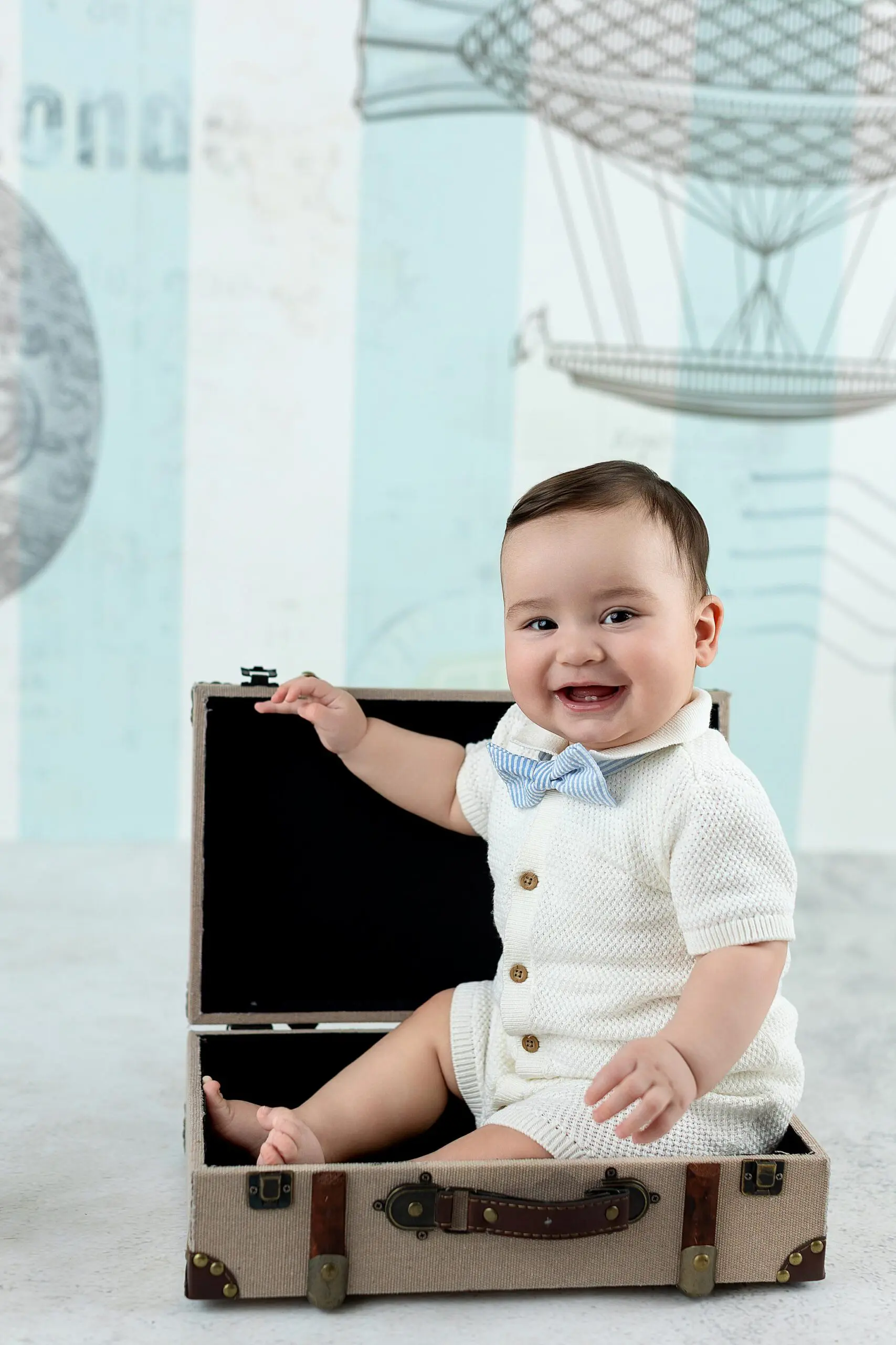 A smiling baby wearing a white outfit and blue bow tie sits inside an open vintage suitcase, with a backdrop featuring hot air balloon illustrations, perfect for charming children portraits.