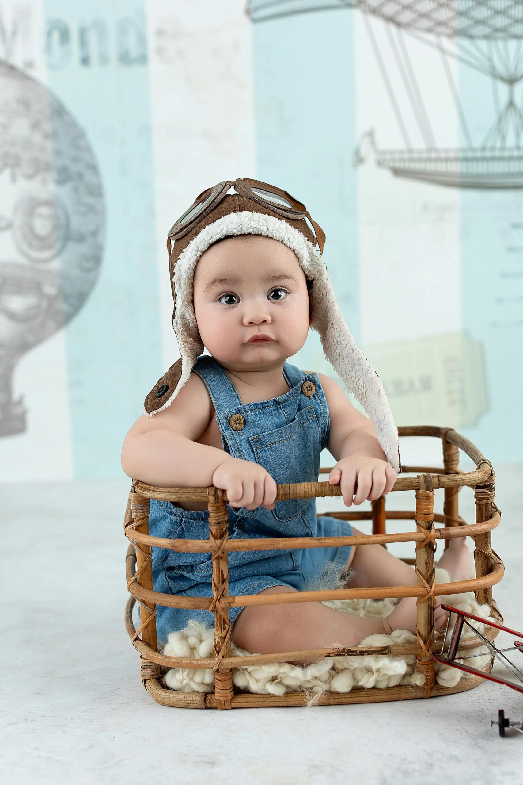 A baby wearing denim overalls and an aviator hat sits in a wicker basket, surrounded by aviation-themed decor, including a drawing of a hot air balloon in the background in Connecticut.
