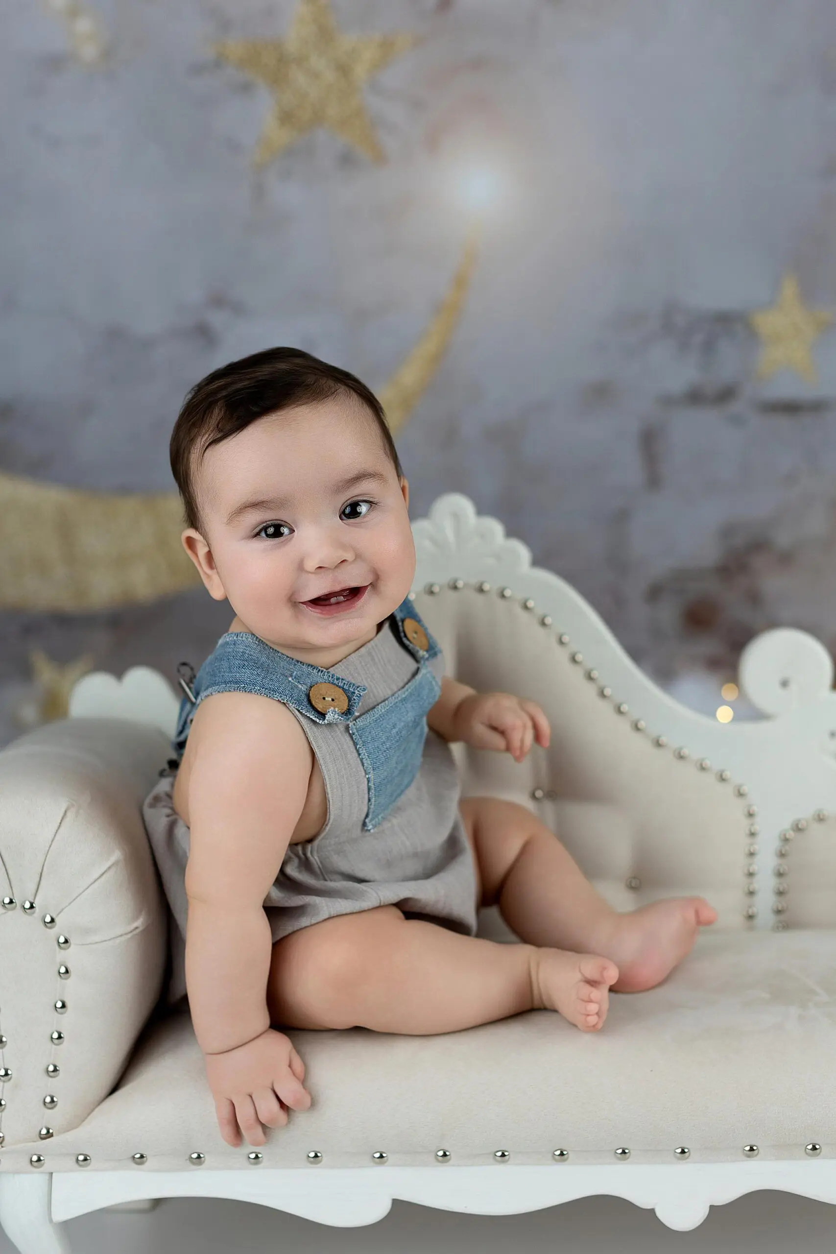 A smiling baby sits on a cream-colored, ornate couch, wearing a gray romper with denim straps. The background features gold stars and a crescent moon on a gray backdrop in a Milford Studio.