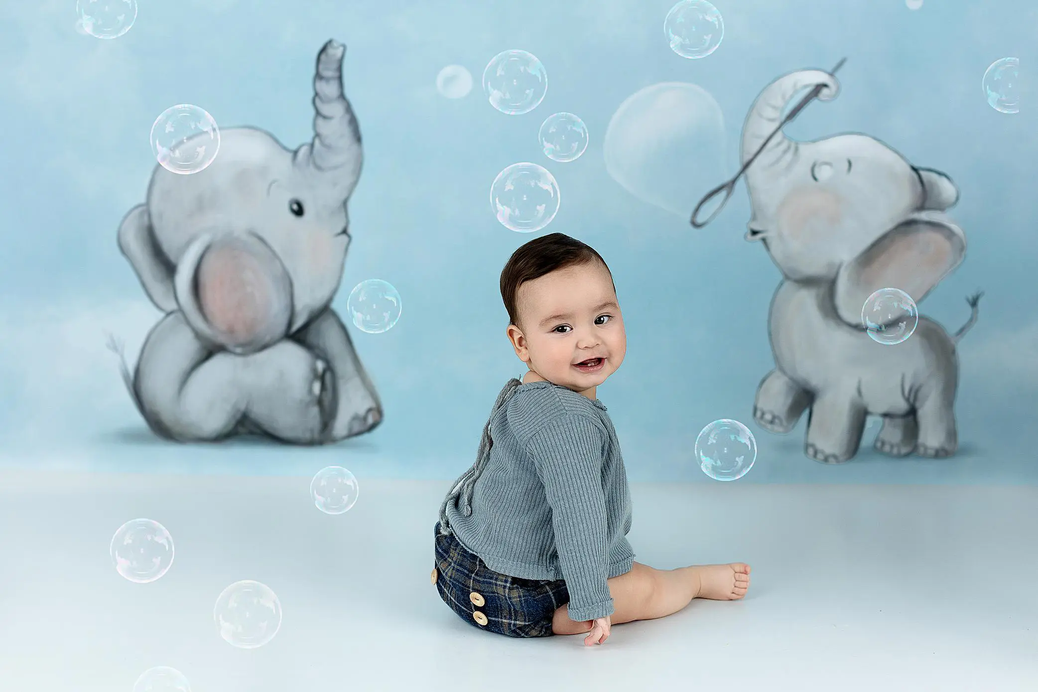 A smiling baby sits on the floor in front of a blue backdrop with cartoon elephants and floating bubbles, wearing a grey sweater and plaid shorts.