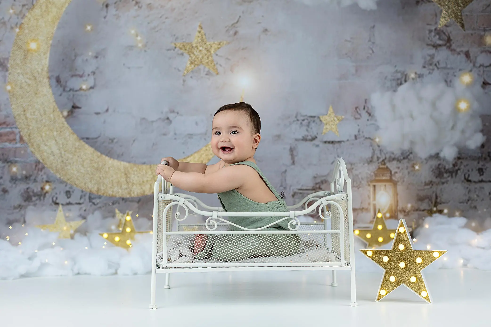 A smiling baby in a green outfit sits on a small white metal bed. The background features gold stars, a crescent moon, clouds, and fairy lights, creating a dreamy, whimsical scene.