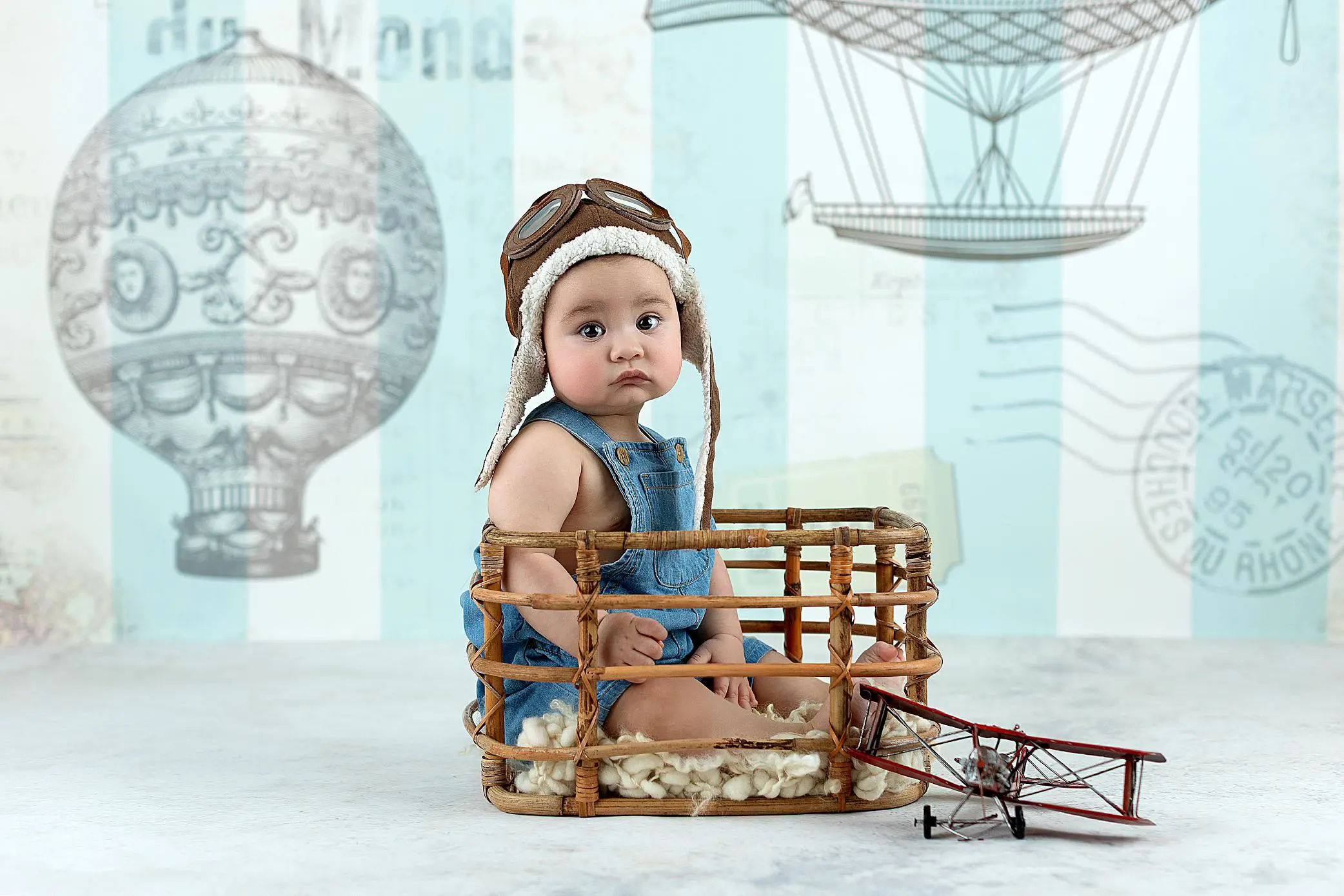 A baby wearing aviator goggles and a pilot hat sits in a wicker basket with wool inside, next to a toy biplane, against a backdrop featuring vintage hot air balloon illustrations.