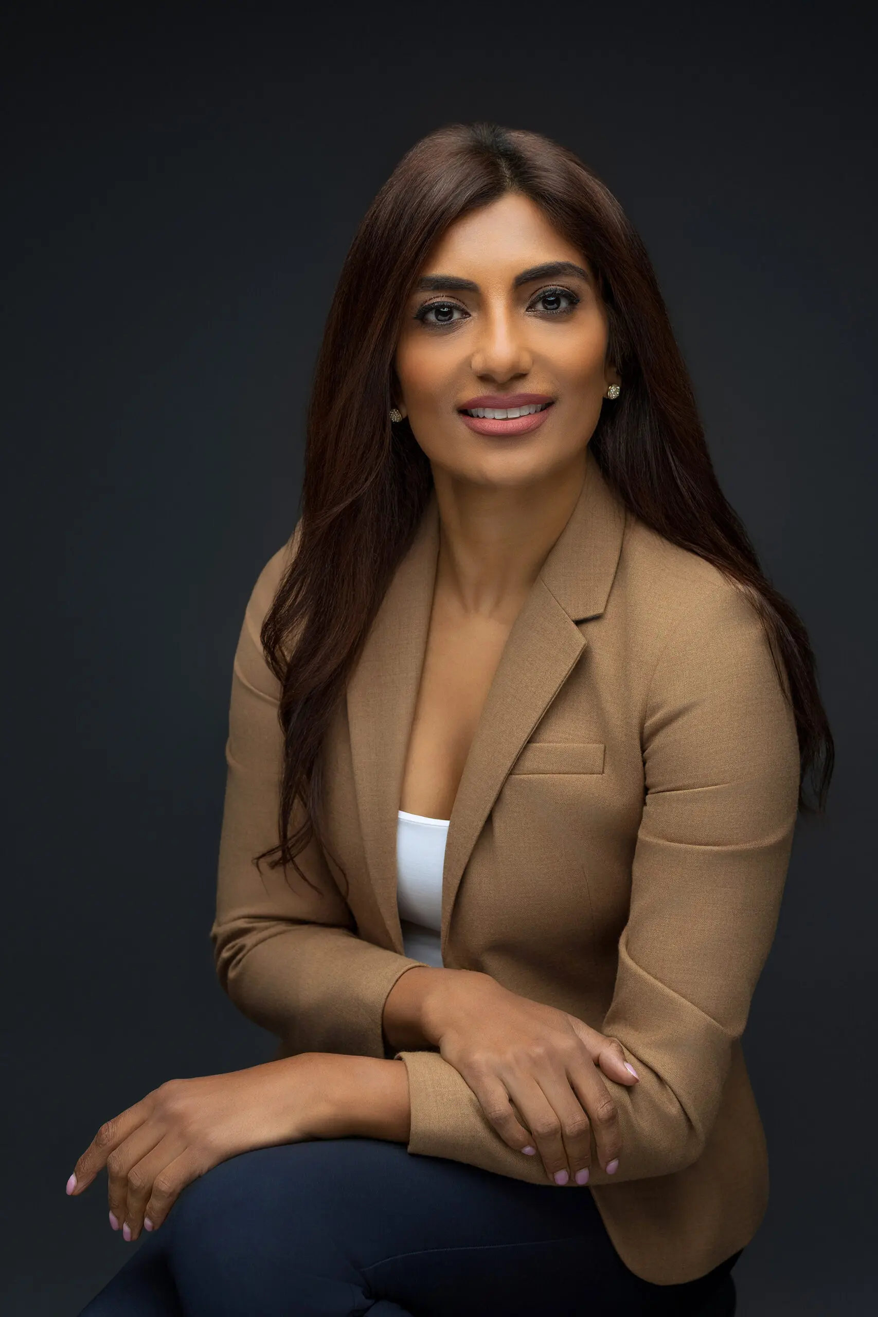 A woman with long dark hair wearing a tan blazer and white top sits against a dark background, smiling with her arms crossed on her lap.