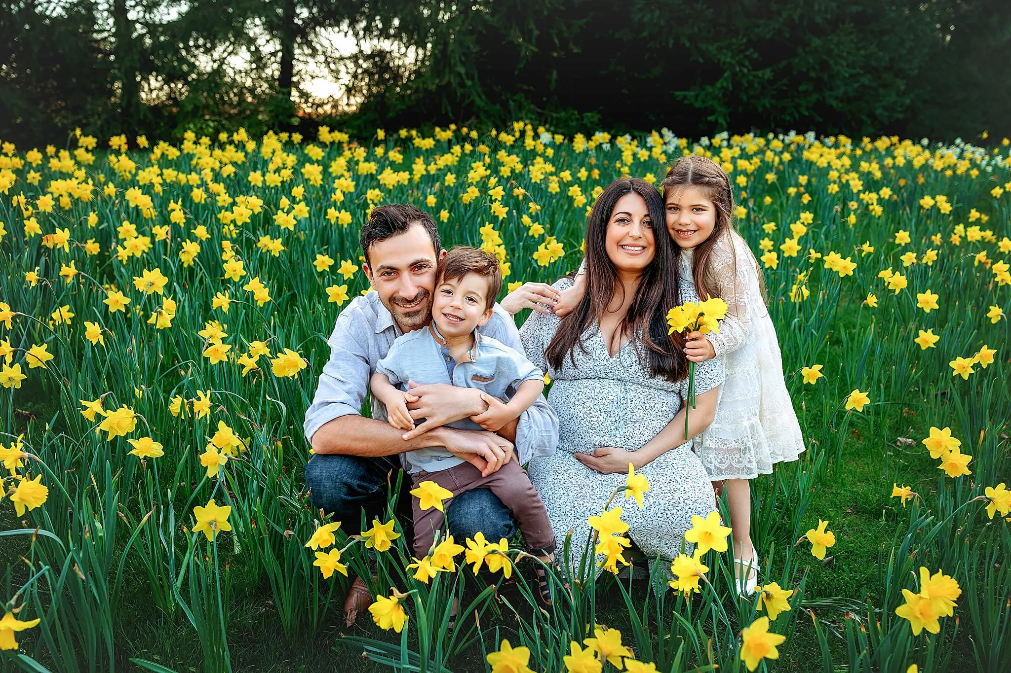 A smiling family of four is kneeling in a vibrant field of yellow daffodils. The man is hugging a young boy, while the woman, visibly pregnant, holds a bouquet. A young girl stands beside them, holding more flowers. Its a sunny day.