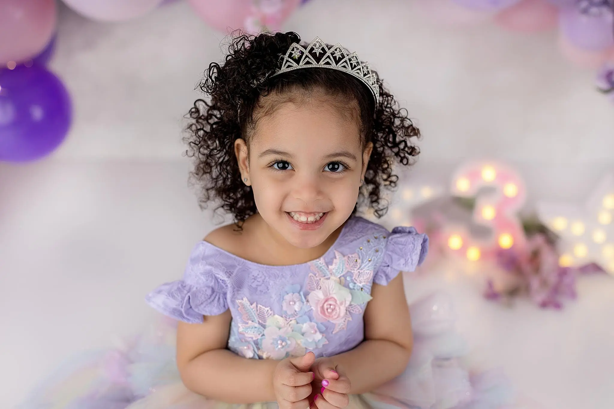 A young girl in a lavender dress adorned with floral details and a tiara smiles warmly. She poses in front of a whimsical backdrop featuring pink and purple balloons alongside illuminated numbers, creating the perfect moment for ct child photos.