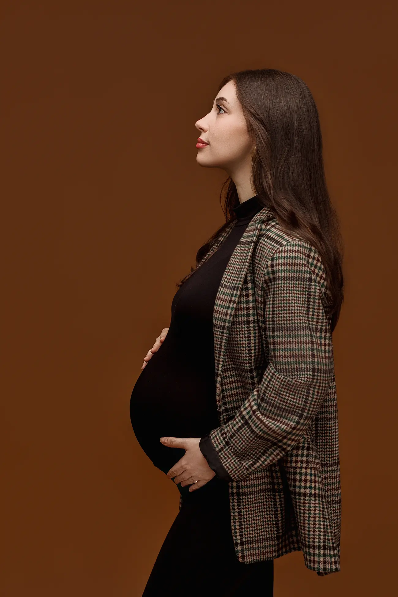A pregnant woman in profile stands against a brown background, wearing a plaid jacket over a black outfit, gently holding her belly and looking upward with a calm expression.