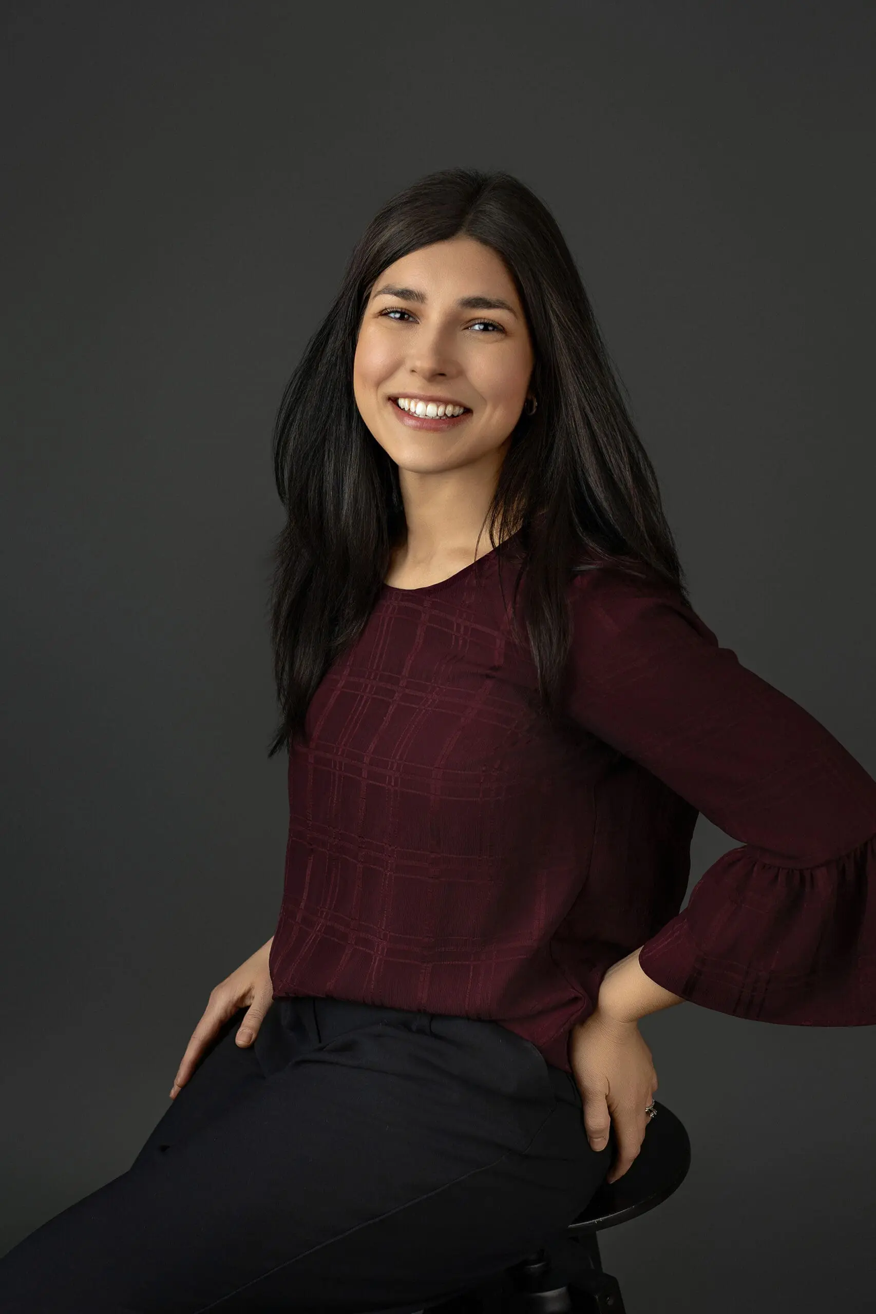 A woman with long dark hair, wearing a maroon blouse and black pants, is sitting on a stool against a dark gray background, smiling with her hands resting on her hips during her studio branding session in Connecticut.