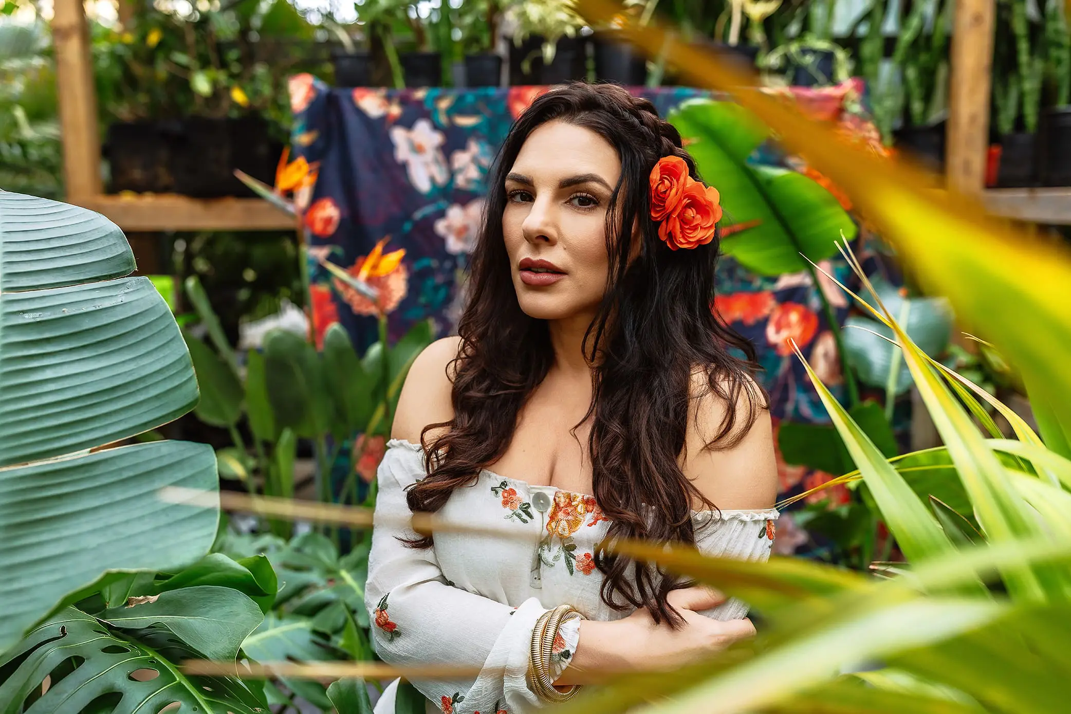 A woman with long dark hair and an orange flower in her hair poses for headshot photos amidst lush green plants. Shes wearing an off-shoulder floral dress, framed by leaves, with a floral fabric backdrop adding to the images vibrant nature.