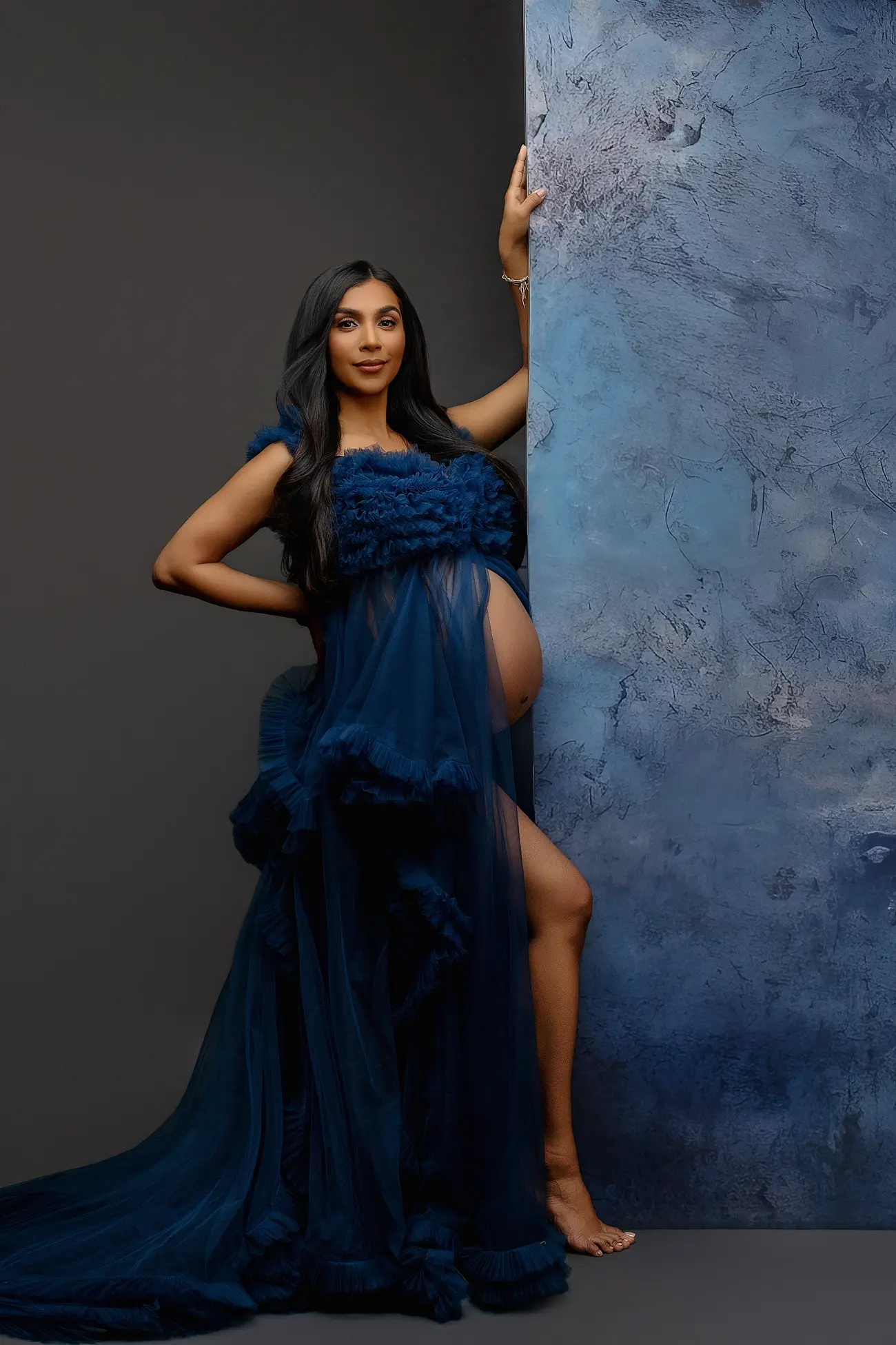 A pregnant woman in a sheer dark blue gown poses barefoot against a textured blue wall, with one hand on her hip and the other resting on the wall, looking confidently at the camera.