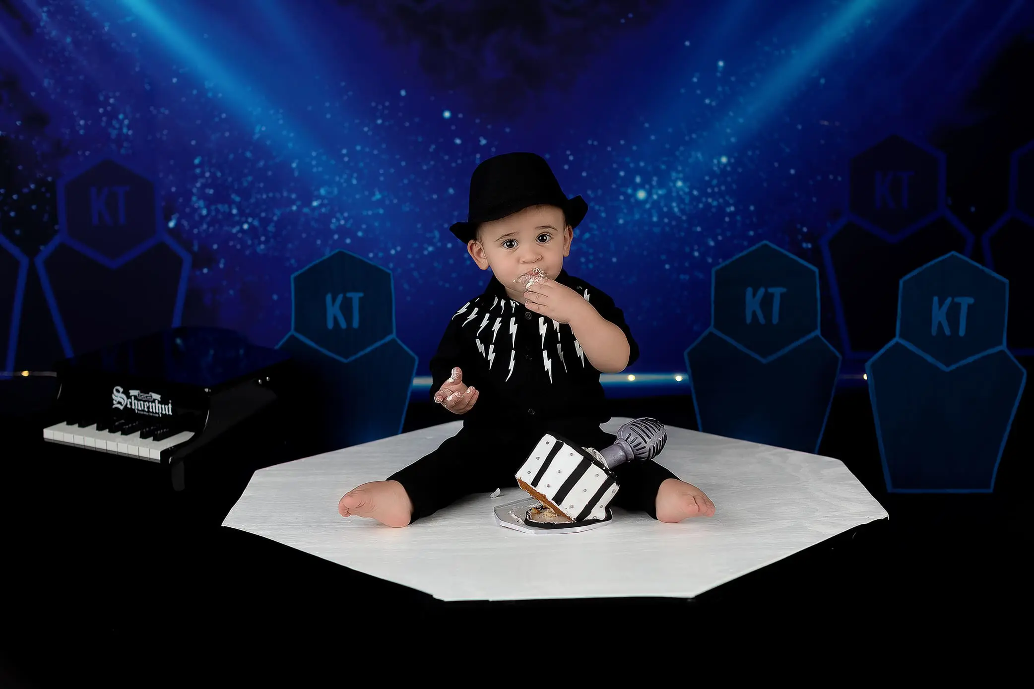 A baby wearing a black outfit with a hat sits on a white surface. The baby is eating cake with frosting on their face, surrounded by blue and black hexagonal decor and a small black piano on the left.
