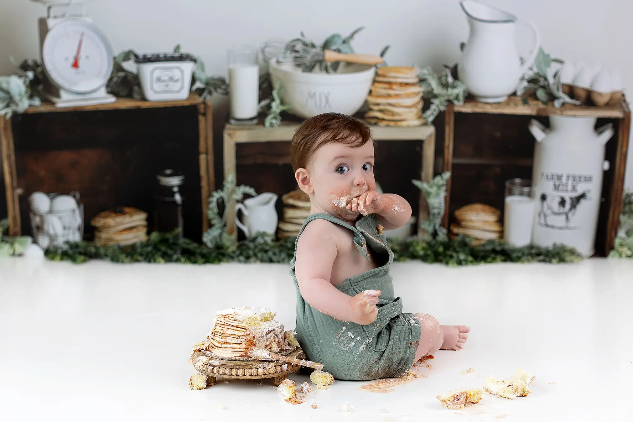A baby wearing a green outfit sits on the floor with a messy face and hands, surrounded by pancakes and frosting. The backdrop includes rustic kitchen decor like scales, pitchers, and crates filled with greenery and kitchen items.