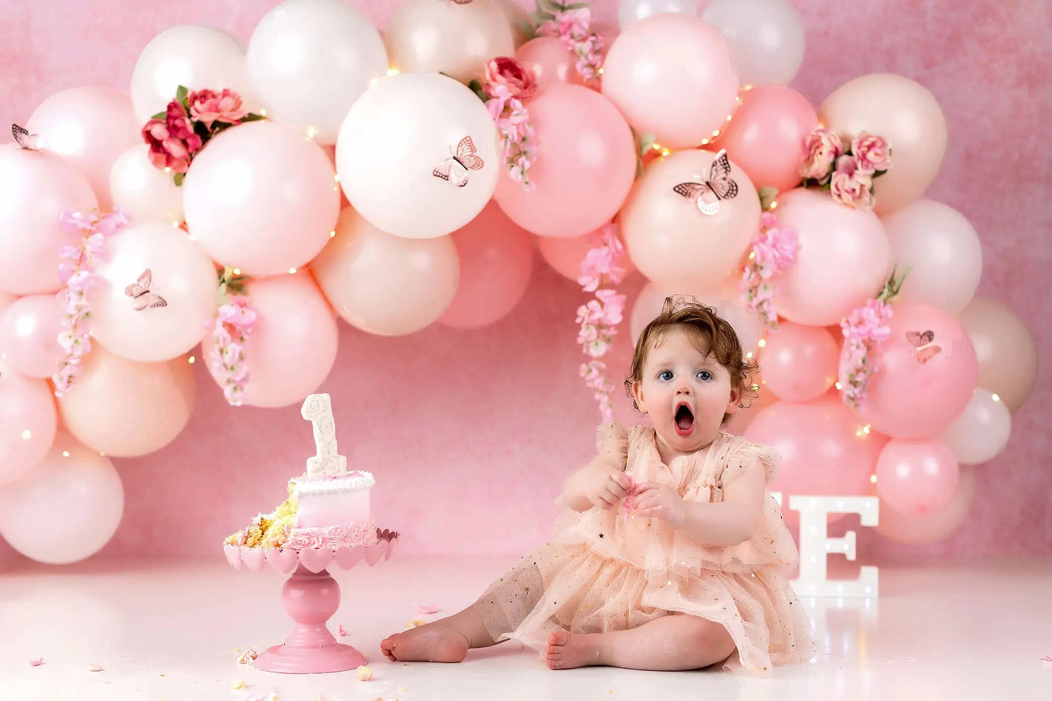 A baby wearing a pink dress sits on the floor, surrounded by pink and white balloons and butterfly decorations. The baby has a surprised expression. A pink cake with a 1 topper is on a nearby stand to celebrate her first Birthday. The background is pink with flowers.
