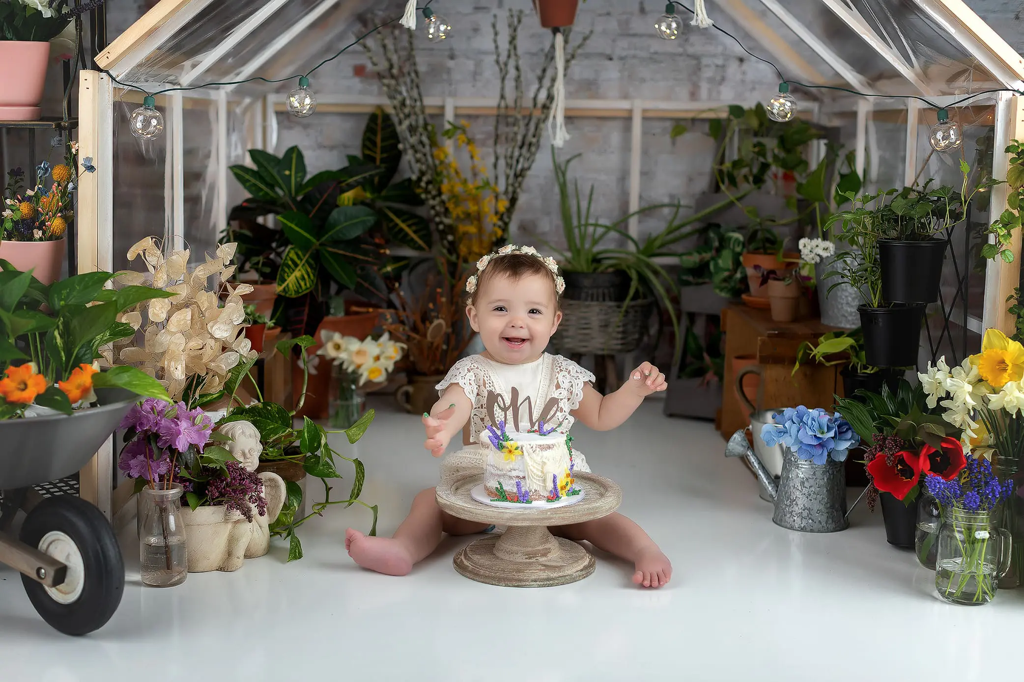 A baby wearing a lace outfit and a floral headband sits on the floor of a greenhouse surrounded by potted plants and flowers. In front of them is a small, colorful cake on a pedestal. The baby is smiling and gesturing with joy.