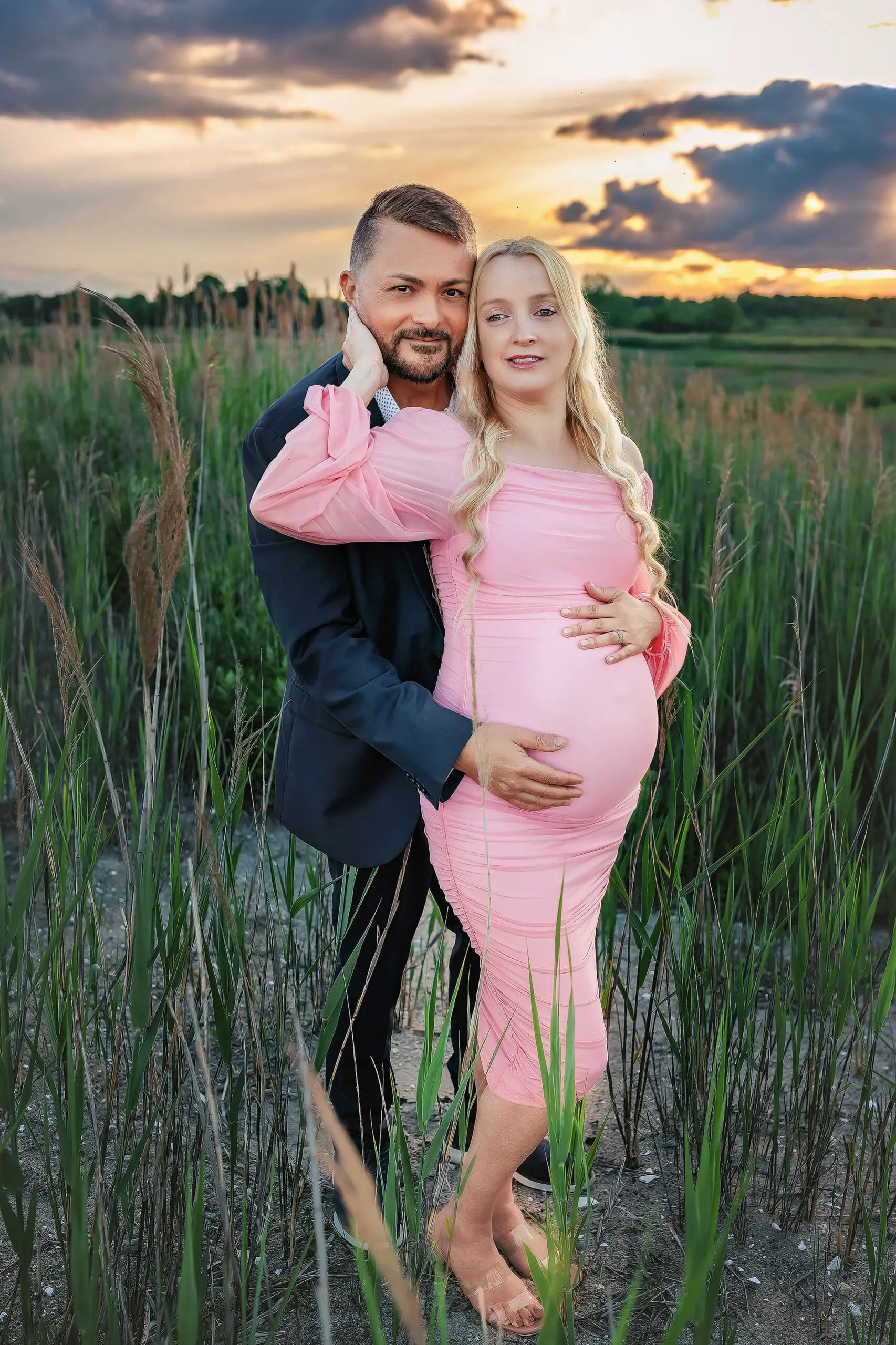 A pregnant couple stands in a grassy field at sunset. The woman, wearing a pink dress, cradles her belly, while the man in a dark suit embraces her. The sky is colorful with clouds and the landscape is lush and serene on a Milford Beach.