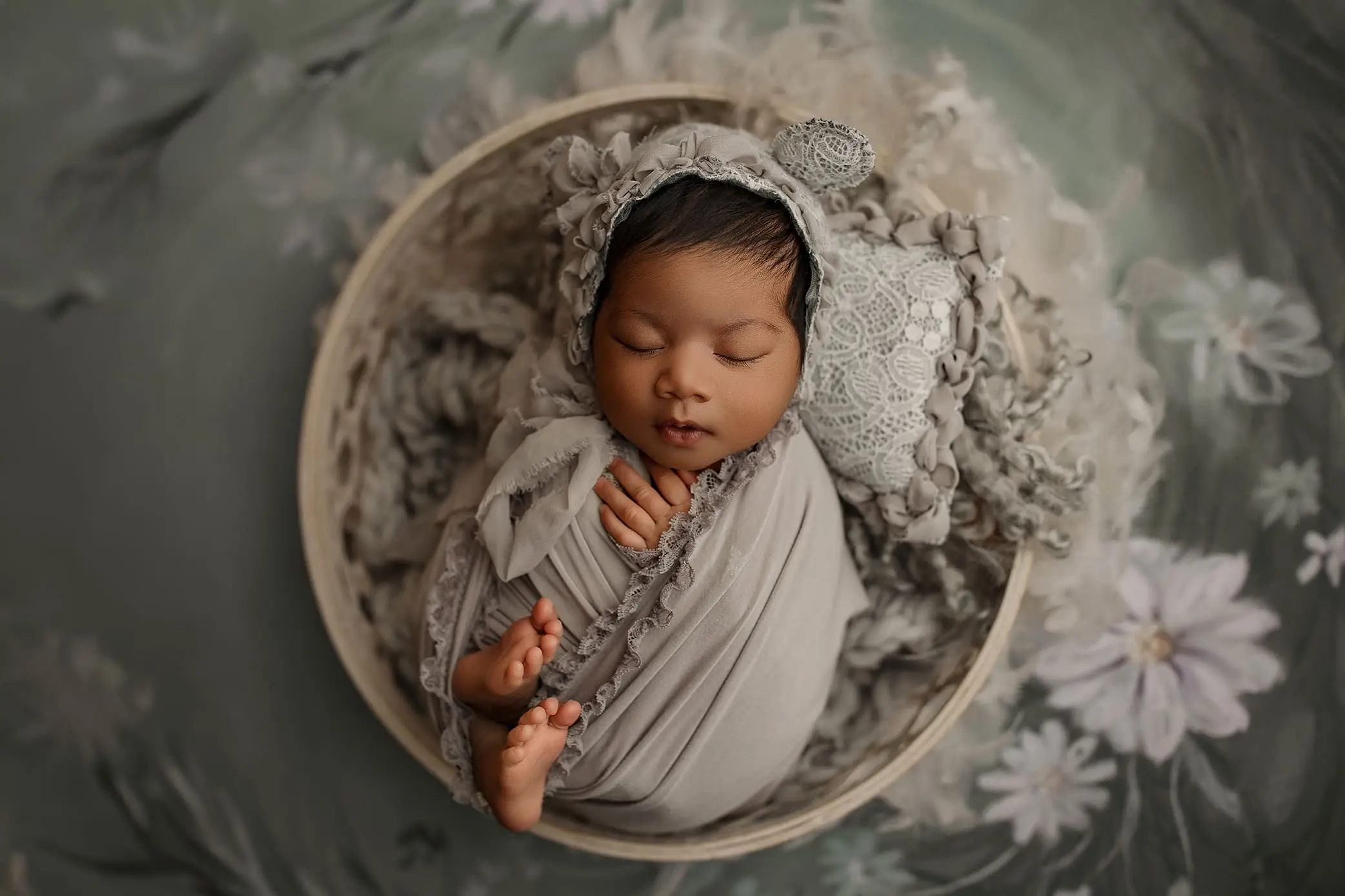 A sleeping baby, wrapped in a soft gray blanket with frilled edges, rests peacefully in a round basket, captured beautifully by a Connecticut newborn photographer. The baby wears a matching lace bonnet. The background features soft floral patterns in muted green and gray tones.