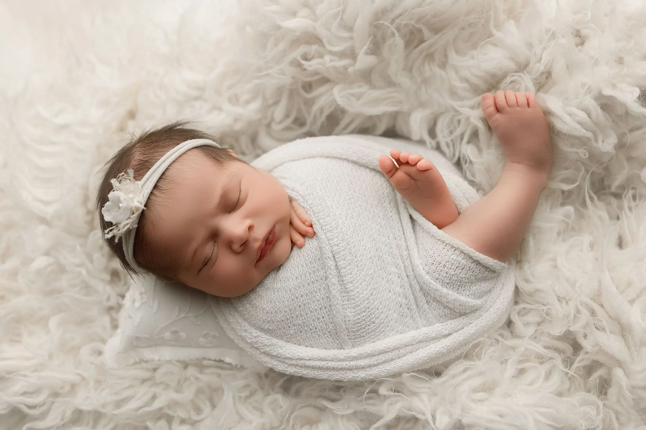 A sleeping baby wrapped in a white blanket lies peacefully on a fluffy, white surface. Captured by a Connecticut newborn photographer, the baby wears a soft headband with a small flower, showcasing an innocent and serene expression.