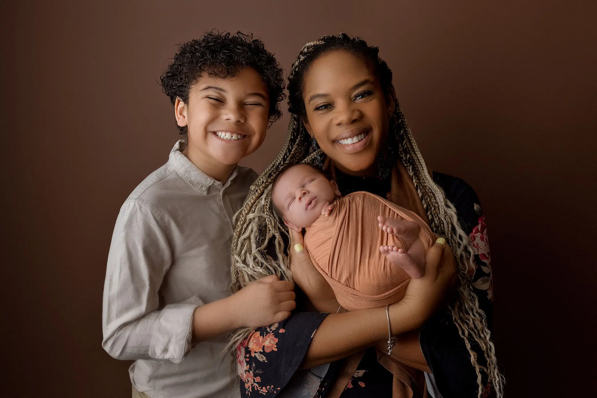A woman with long braided hair smiles while holding a newborn wrapped in a warm, orange cloth, seemingly capturing the essence of newborn photos CT. Beside her, a young boy with curly hair grins widely. They stand against a plain brown background, exuding warmth and happiness.