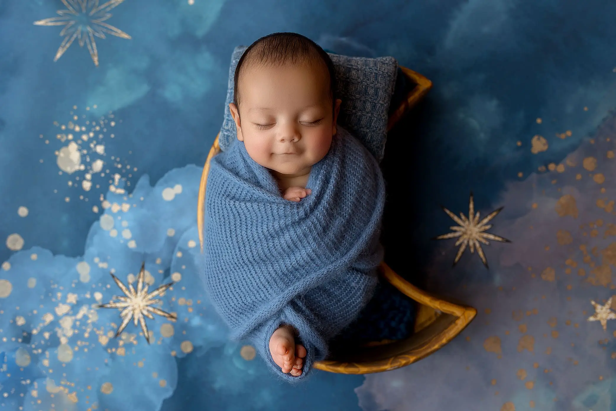A newborn, wrapped in a soft blue blanket, sleeps peacefully in a crescent-shaped prop against a dreamy, starry blue background. The babys foot peeks out from the blanket, surrounded by gold and white star-like decorations—a perfect setting for newborn photos.