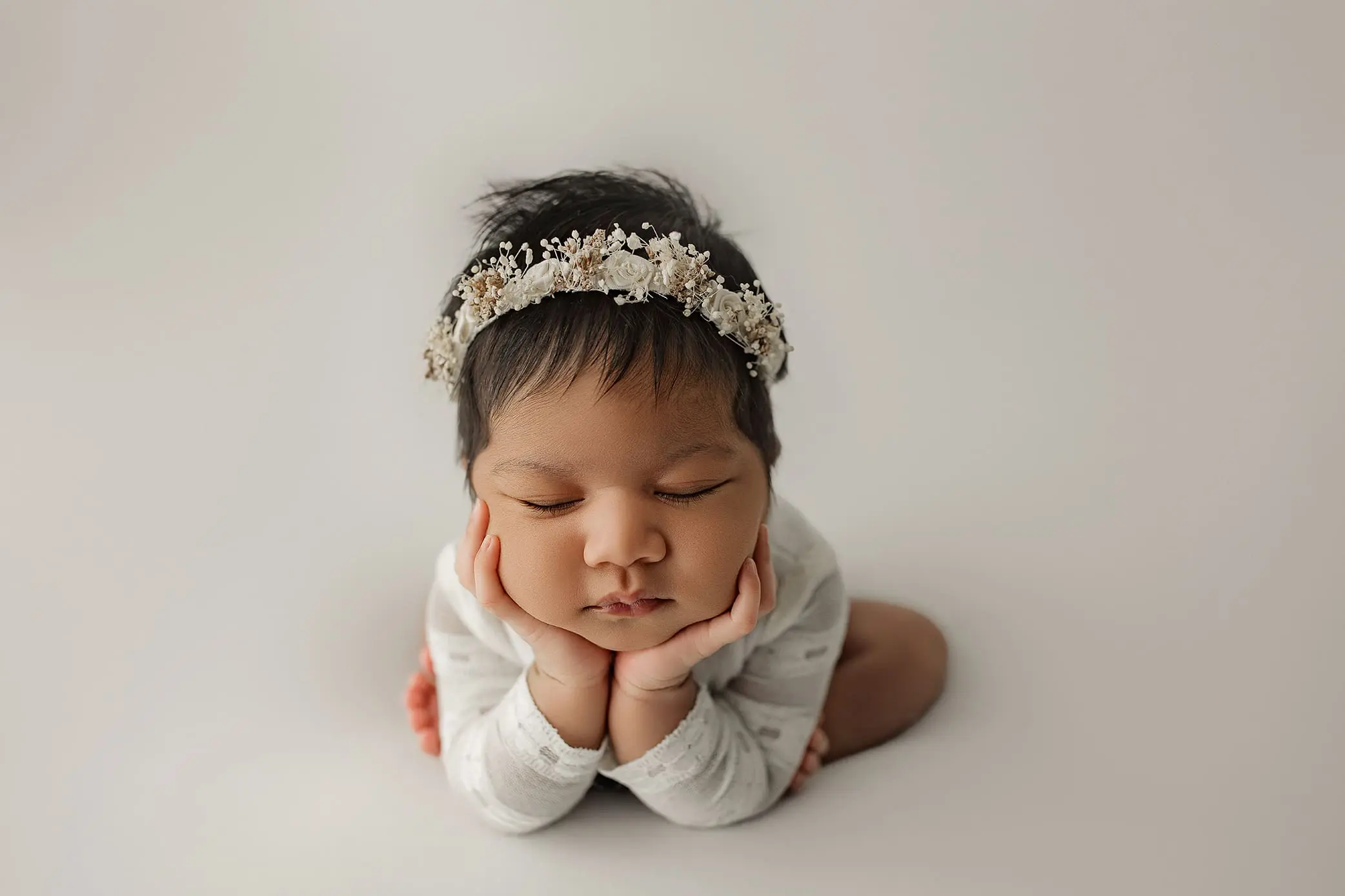 A newborn in Connecticut with closed eyes rests their head on their hands. They wear a delicate floral headband and a white outfit, set against a soft, neutral background.