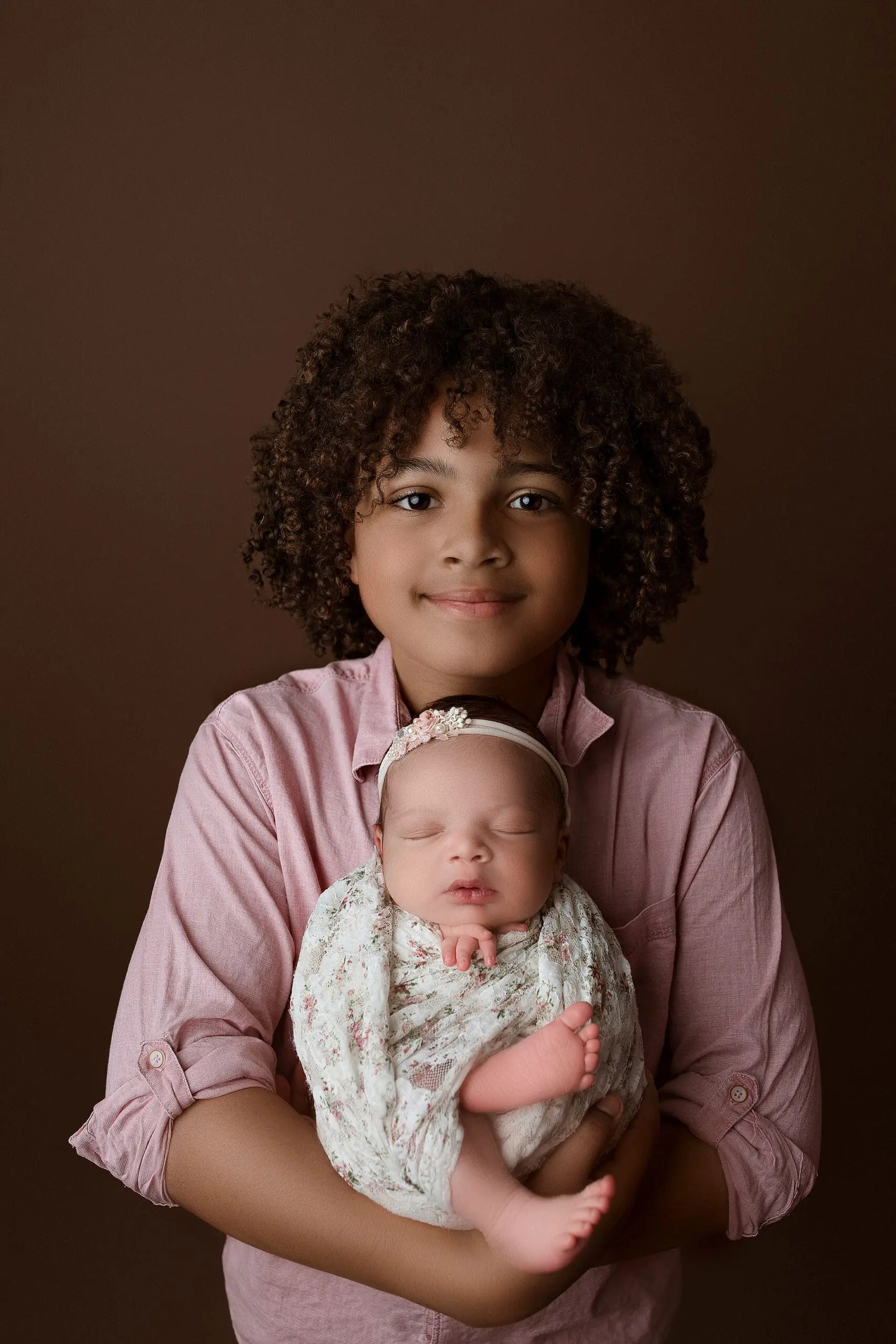 A child with curly hair, wearing a pink shirt, gently holds a sleeping baby wrapped in a floral blanket against a brown background. The baby is adorned with a matching floral headband.