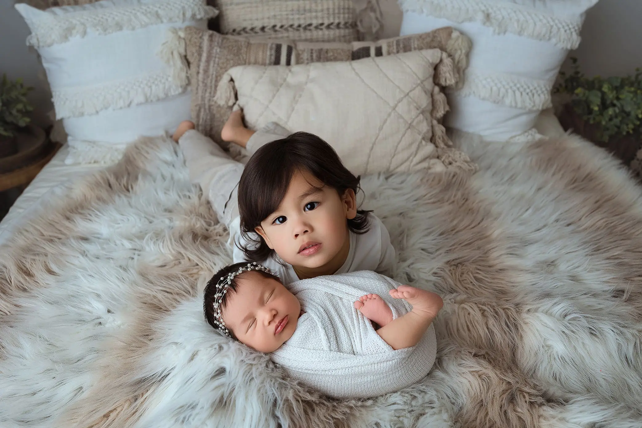A young child lies on a fluffy, cream-colored blanket, next to a sleeping baby wrapped in a white swaddle with a headband. Capturing the essence of newborn photos in Connecticut, they rest on a bed with textured pillows, creating a cozy and serene setting.