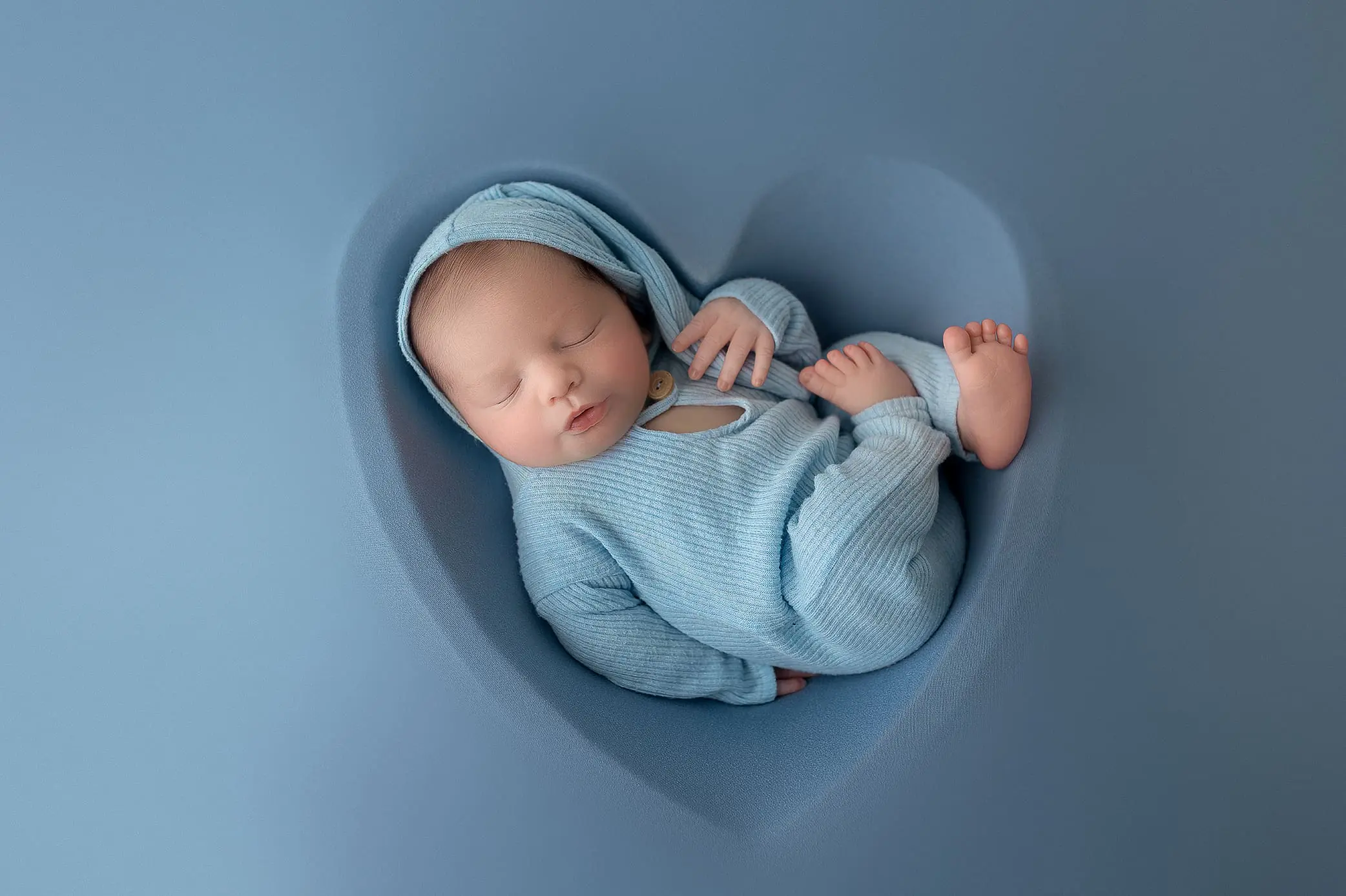 A sleeping baby dressed in a blue outfit and cap, nestled comfortably within a heart-shaped cushion against a soft blue background. The babys peaceful expression and tiny feet are visible.
