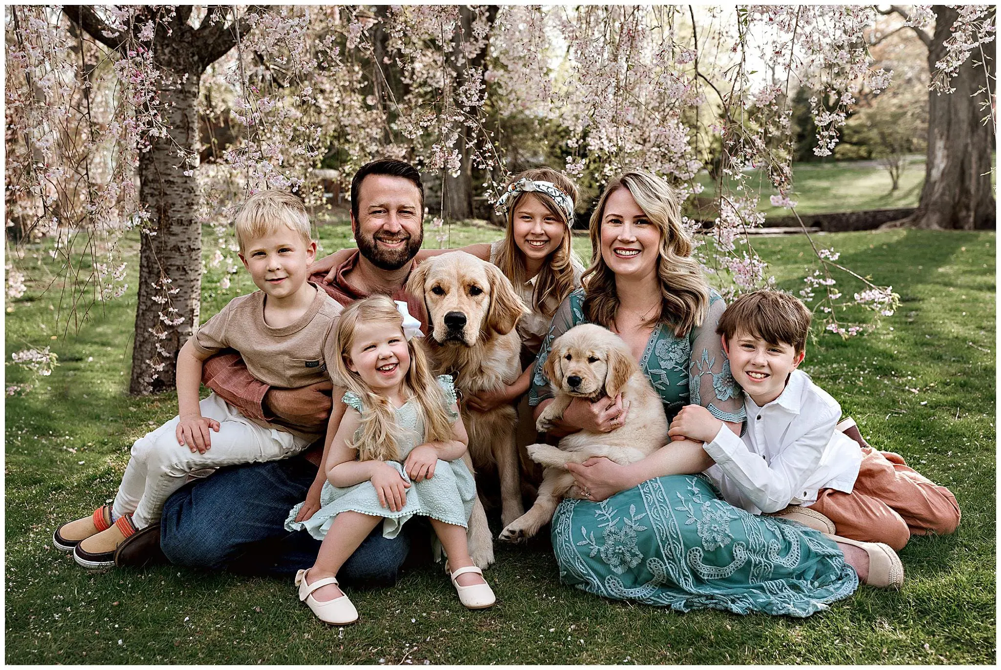 A family of five, including three children, poses outdoors under blooming trees in Milford, Connecticut, with two golden retrievers. The group is sitting on the grass and smiling at the camera, dressed casually in pastel colors.