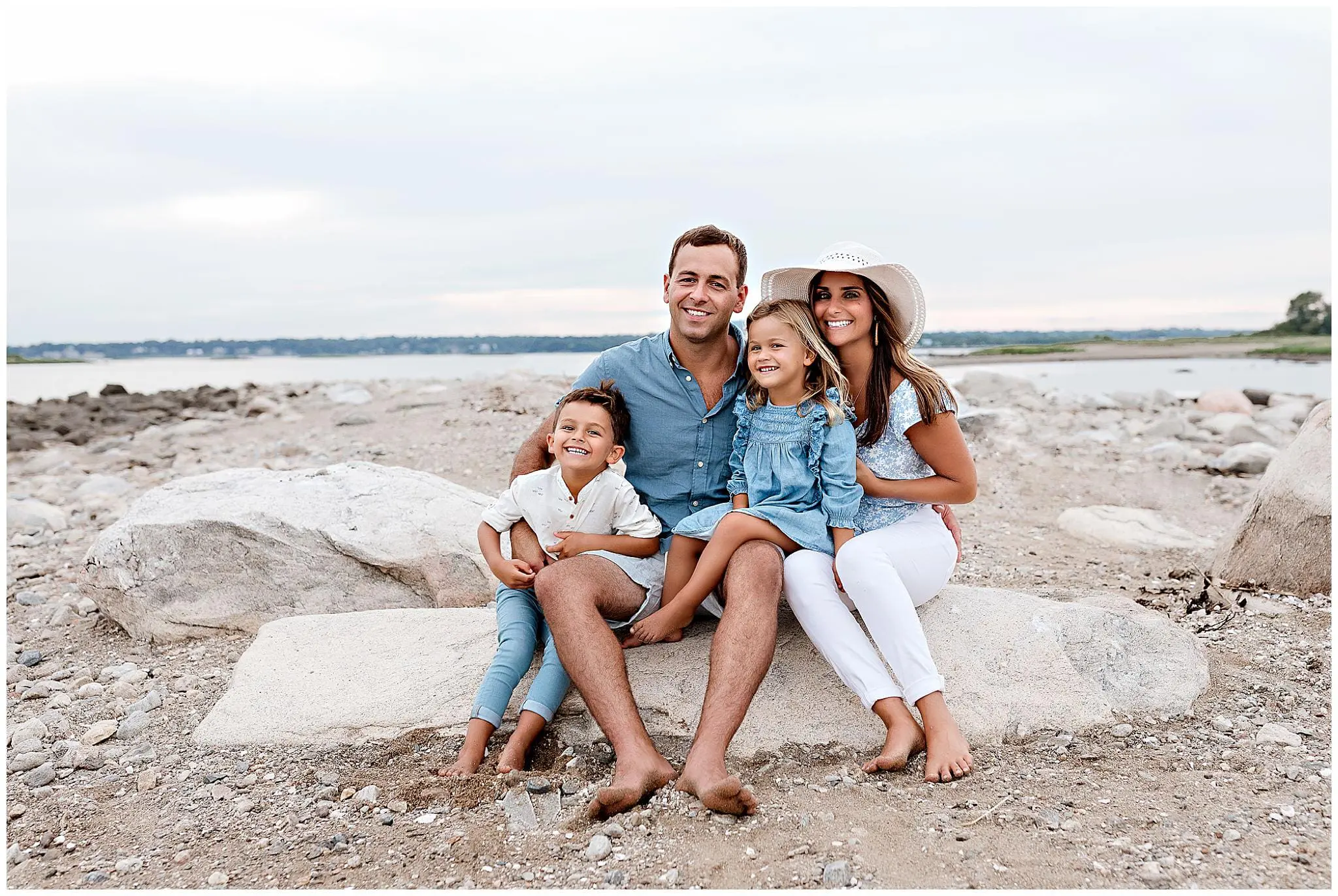 A family of four sits on a rock at the beach, smiling. The father and mother are with their young son and daughter. The scene is set against a backdrop of sand, rocks, and a sea under a cloudy sky. The mother wears a sunhat.