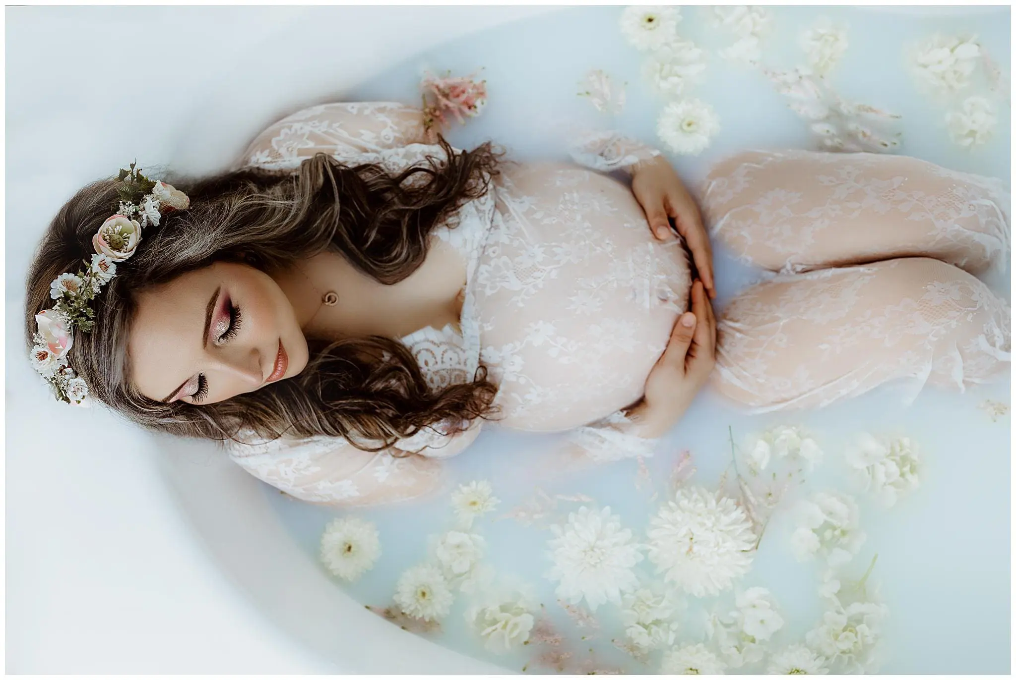 A graceful maternity scene unfolds as a pregnant woman in a sheer lace dress relaxes in a milky bath. Cradling her belly with both hands, she is surrounded by white flowers and pink roses, while a floral headband adorns her closed eyes, evoking tranquility akin to the serenity of Milford.