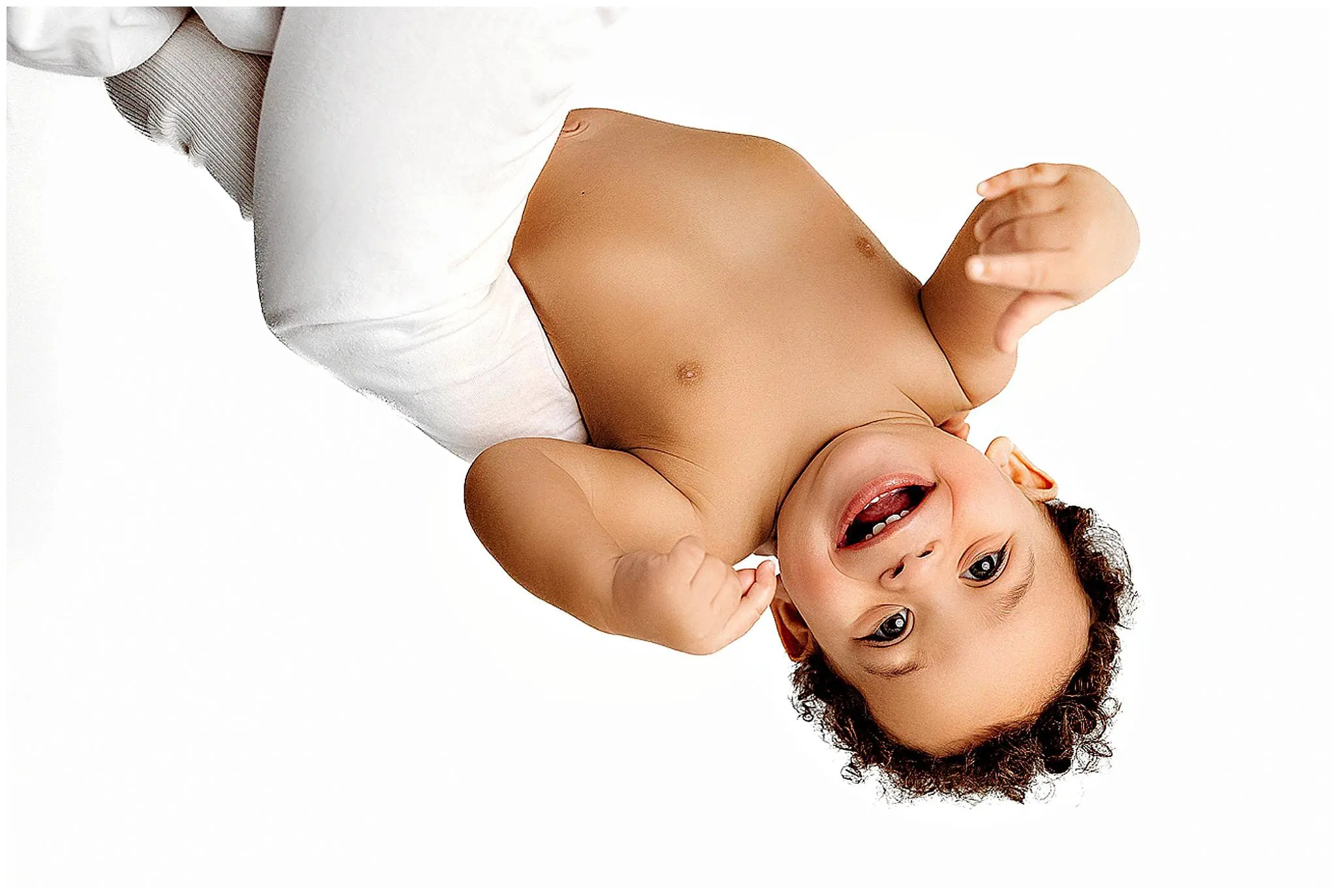 A curly-haired baby from Milford, Connecticut is held upside down and smiling, wearing only a diaper. The plain white background perfectly highlights the babys joyful expression.