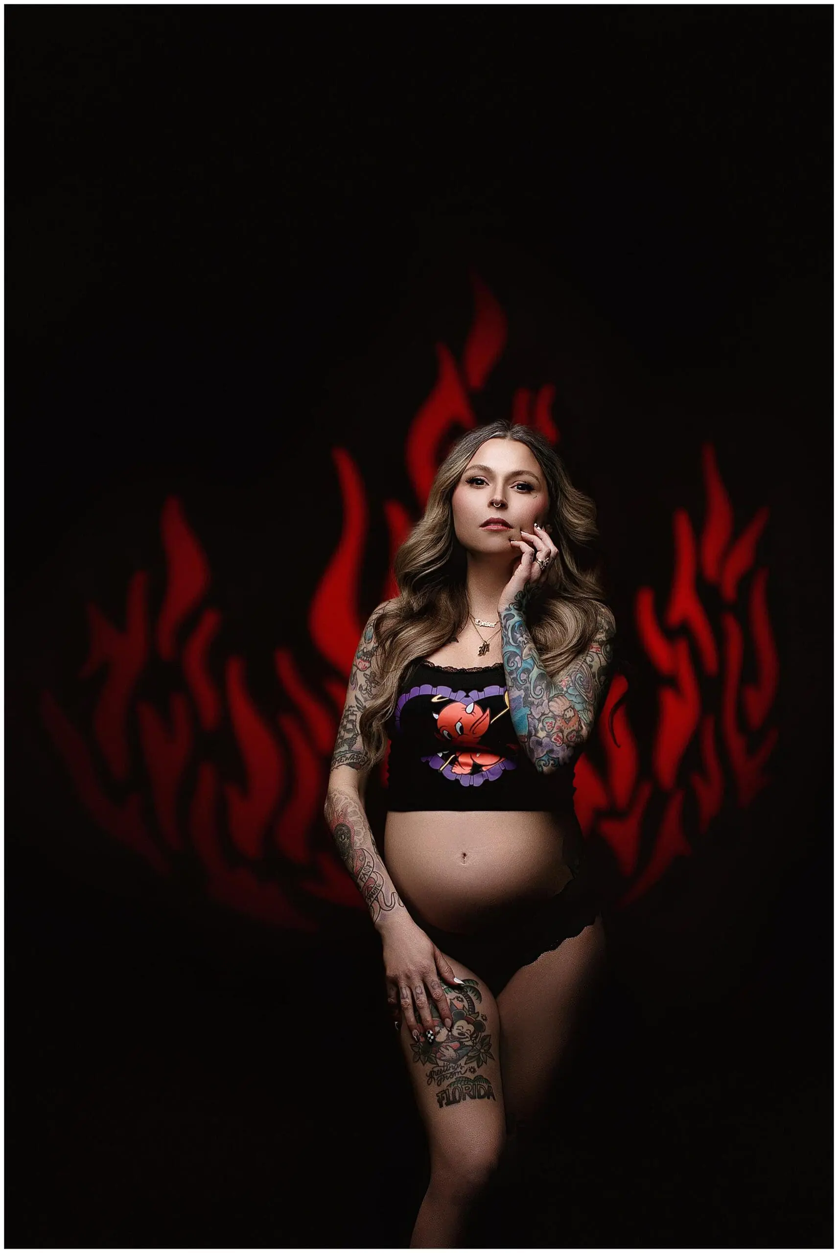A pregnant woman with long, wavy hair and extensive tattoos poses confidently, embracing her Milford maternity style. She wears a black top with a design and matching bottoms. A red flame-like pattern dances in the background against a dark backdrop.