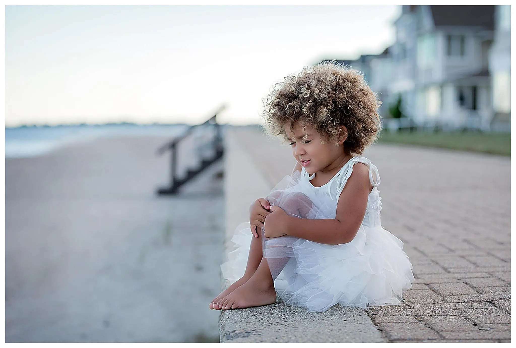 A young child with curly hair sits on a paved ledge by Milford Beach, wearing a white, fluffy dress. Barefoot and smiling gently as they look down, the backdrop of houses and sand blurs softly behind.