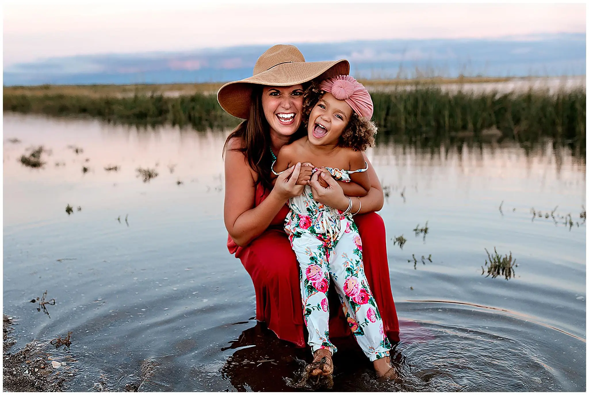 A woman in a wide-brimmed hat and red dress kneels by a lake, smiling with a young girl in colorful floral clothing on her lap. Capturing this moment of joy with Milford Photography, the scene showcases their infectious smiles against a backdrop of grass and shimmering water.