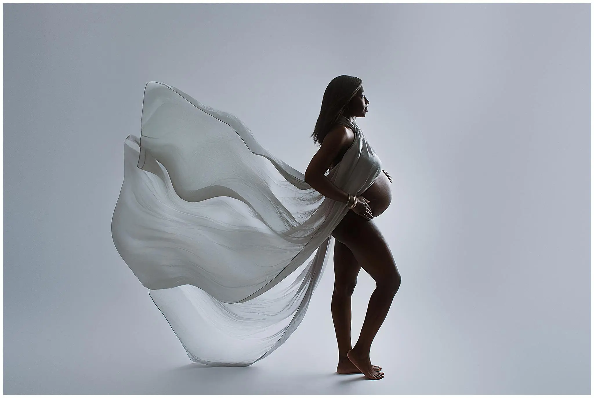 Pregnant woman in a flowing, translucent gown, standing in profile against a neutral background. She holds her belly with one hand, and the fabric elegantly billows behind her, creating a sense of movement.