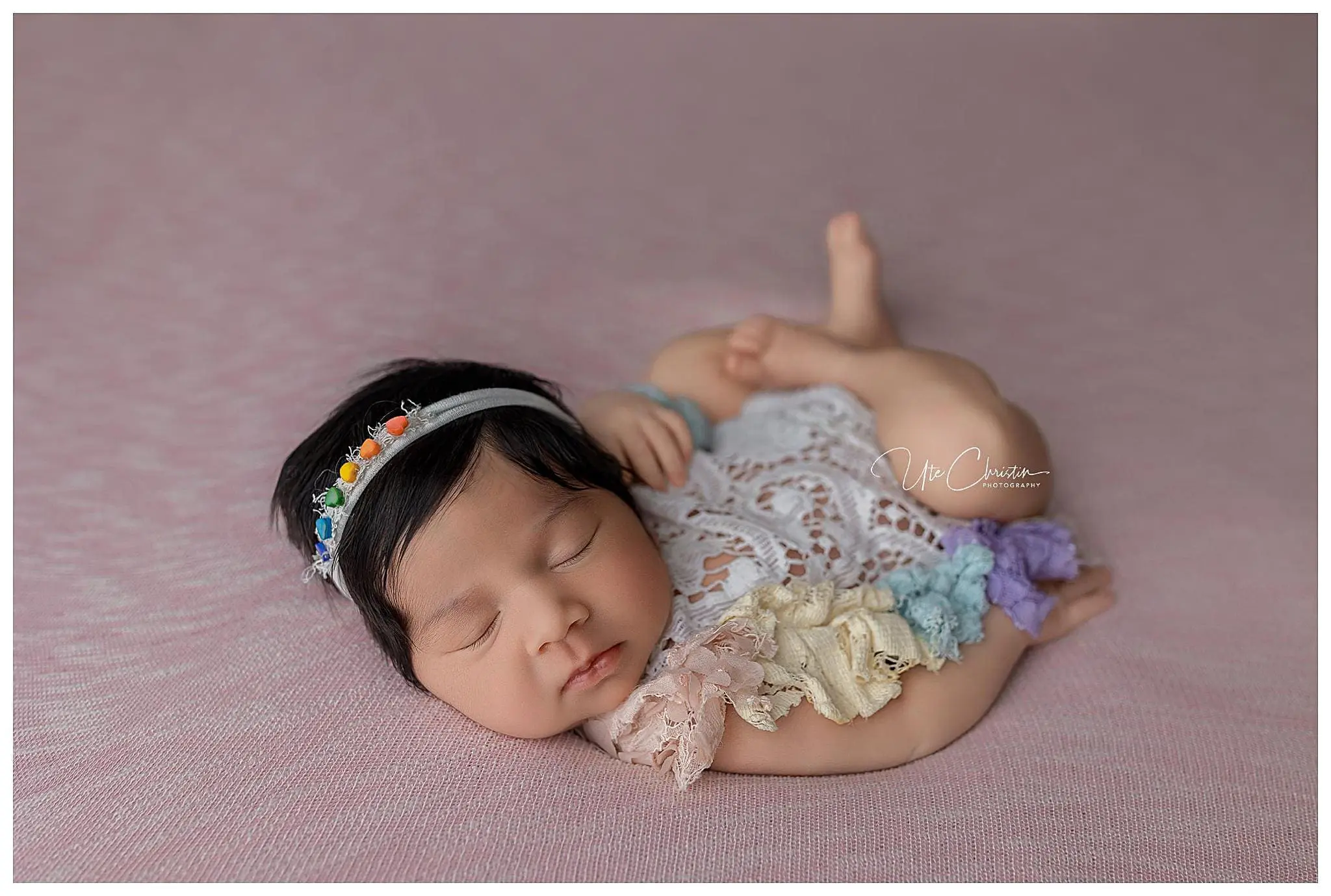A sleeping baby with dark hair lies on a soft pink blanket, wearing a lace outfit adorned with colorful flowers. Their headband features tiny multicolored blooms, procured from one of the charming baby stores in Connecticut. The babys hands are gently placed under their head.