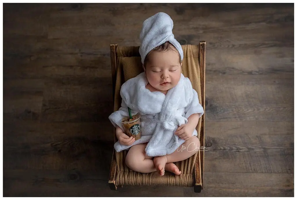 A baby in a white robe and towel turban sleeps peacefully in a small hammock, holding a miniature coffee cup with a green straw. This delightful scene could inspire displays at baby stores in Connecticut. The warm wooden floor adds a cozy touch to this charming moment.