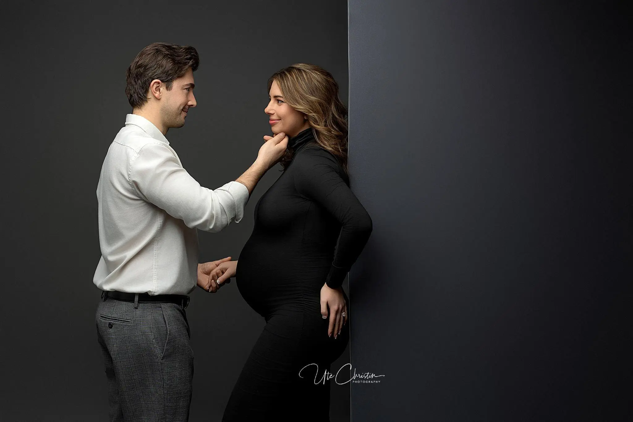 A man gently touches a pregnant womans chin as they stand together outside one of the renowned birthing centers in Connecticut. The woman, wearing a black dress, leans against a dark wall while the man, in his white shirt and grey pants, shares a look of contentment and connection with her.
