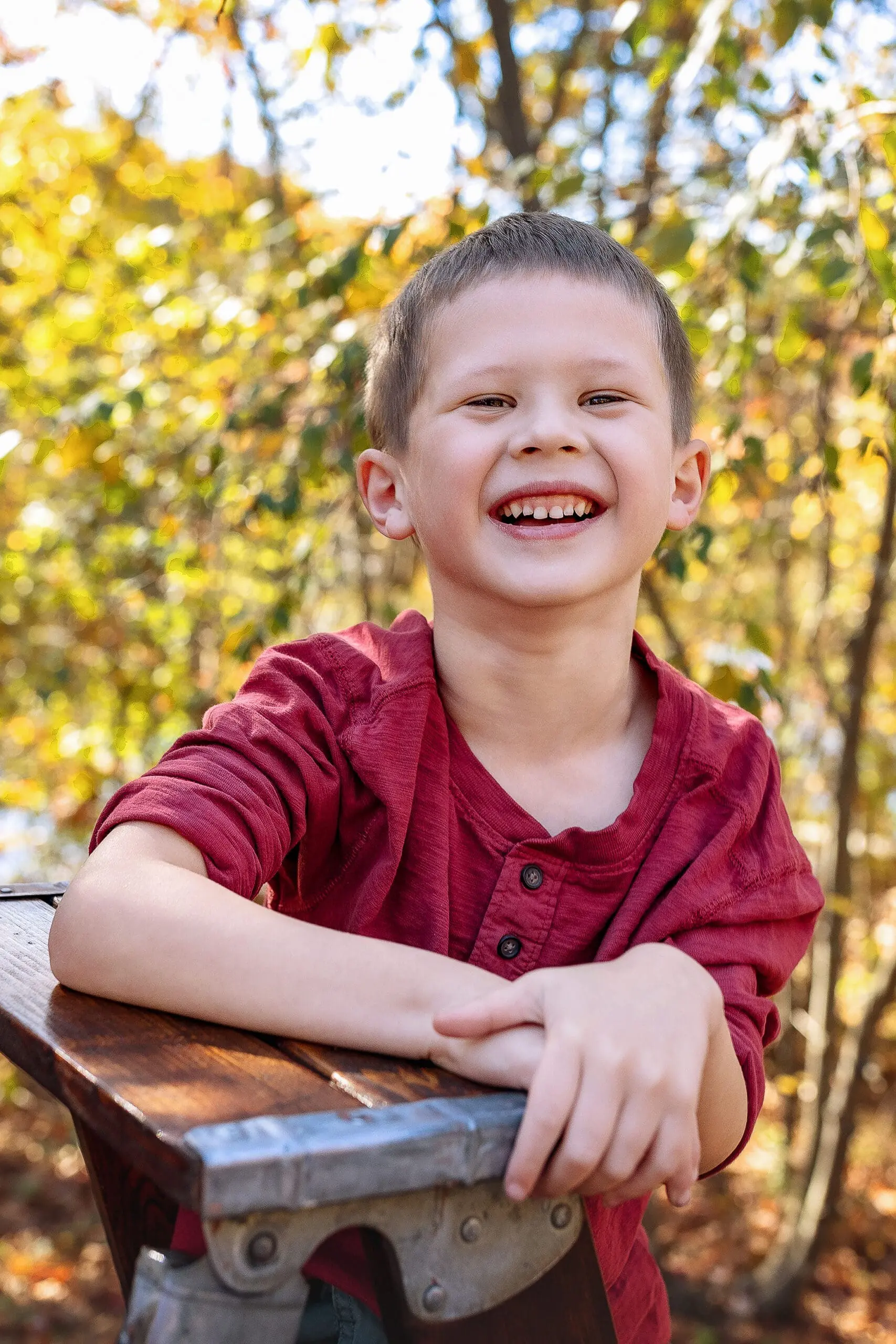 A young boy with short brown hair, wearing a red shirt, smiles brightly while leaning on a wooden bench. The background features blurred trees with autumn leaves in warm colors.
