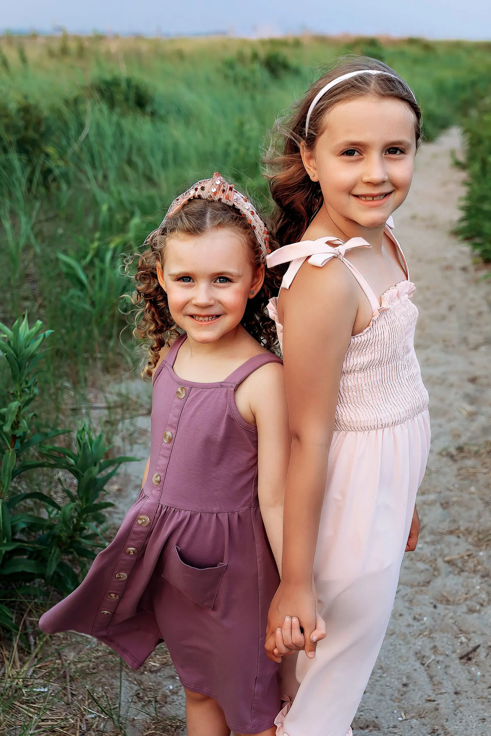 Two young girls stand hand in hand on a sandy path surrounded by greenery. Both are smiling and wear summer dresses, with one in a mauve dress and the other in a pink dress. Their hair is adorned with headbands. The setting is serene and natural.