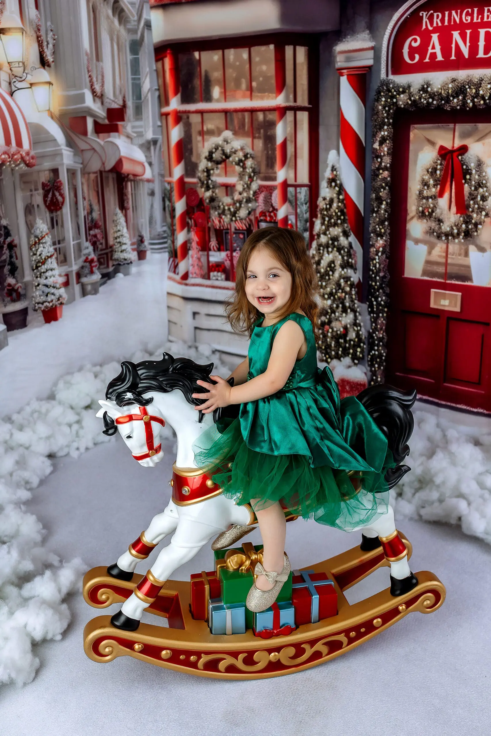 A young girl in a green dress joyfully rides a decorative rocking horse surrounded by festive Christmas decor. The background includes snow, holiday wreaths, and a “Candy” shop entrance, exuding a cheerful seasonal atmosphere in a Connecticut studio photographing holiday minis.