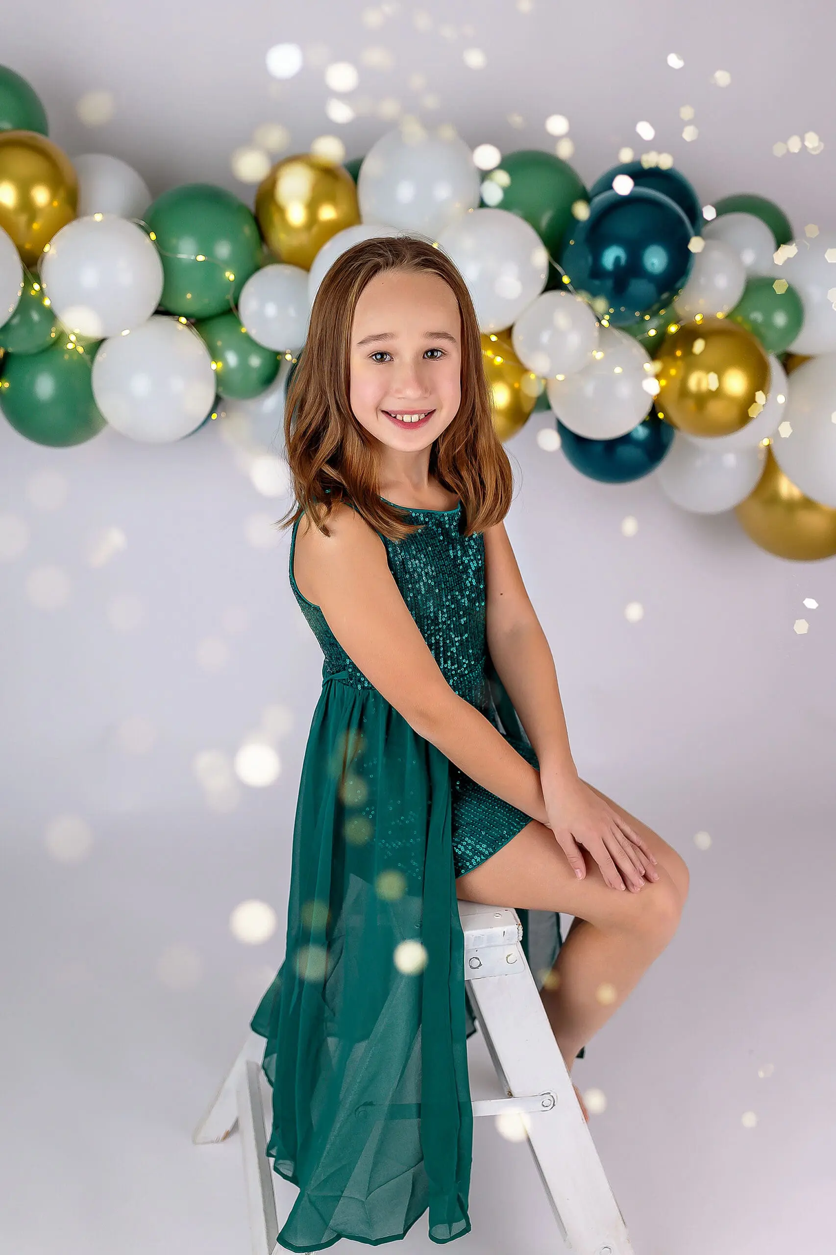 A young girl in a green dress sits on a white ladder, smiling. She is surrounded by a festive balloon arch in white, gold, and green. Soft glowing lights are scattered throughout the image, adding a magical touch to celebrating her 10th birthday with a CT studio session.
