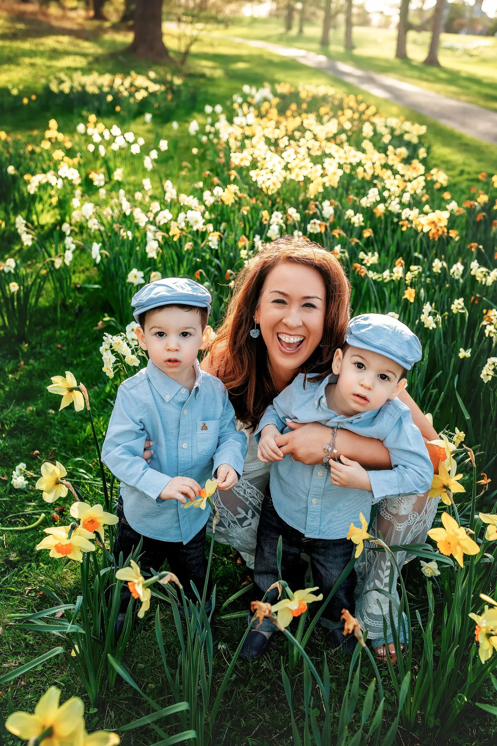 A woman is crouching and smiling, with her arms around two young boys in matching blue shirts and caps, surrounded by blooming daffodils in a sunny park in Connecticut.
