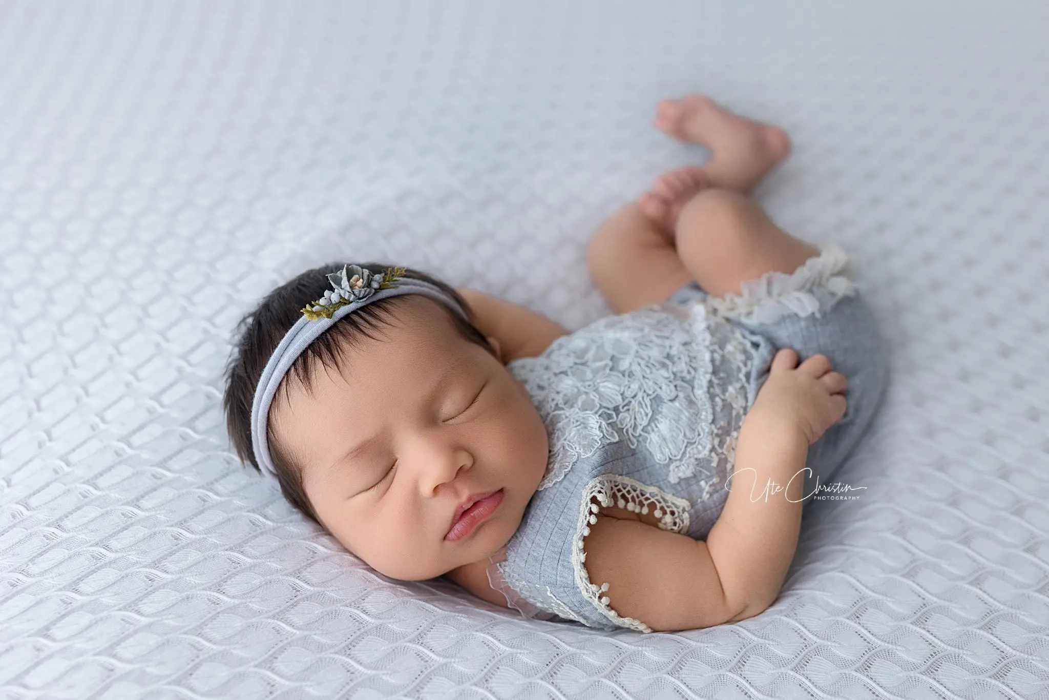 A peaceful sleeping baby, gently cradled by the care of doulas in CT, wears a light blue lace outfit and a floral headband. The baby rests on a textured white blanket, with legs slightly bent and arms softly by their side.