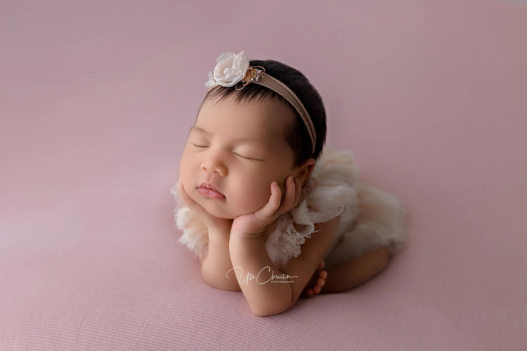 Against a soft pink background, a sleeping baby in white rests peacefully, their head on tiny hands. A flower-adorned headband adds charm to the serene scene. This delicate moment captures the tranquility that Doulas In CT help foster for new families and their precious little ones.
