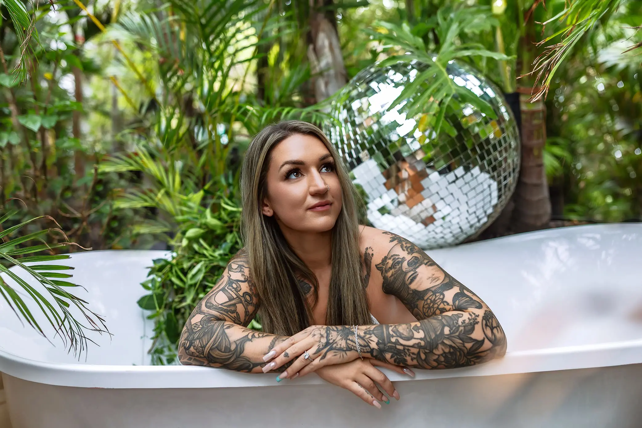 A woman with long hair and extensive tattoos relaxes in a white bathtub surrounded by lush green plants. A large disco ball is partially visible in the background, adding a reflective element to the tropical setting created in a greenhouse in Connecticut.