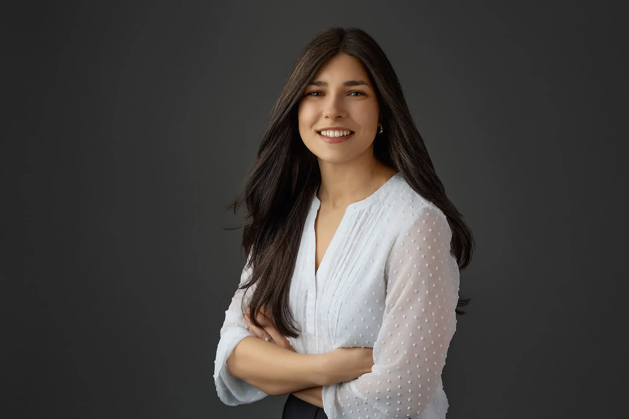 A woman with long dark hair is smiling and looking at the camera. She is wearing a white blouse with textured sleeves and has her arms crossed in front of her. The background is a plain dark gray picked out for her headshot photo session in Milford