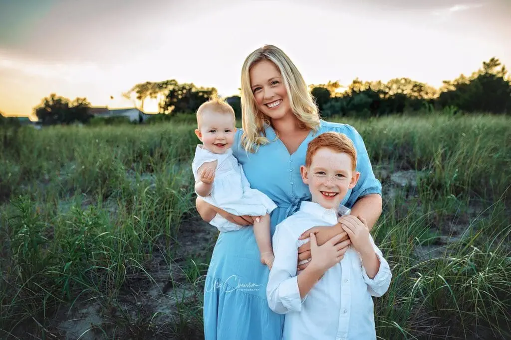 A smiling woman in a blue dress stands outdoors with a young boy and a baby, both in white, during sunset. They are surrounded by green grass and trees—a warm, cheerful family scene perfect for Pediatric Dentists In Milford CT.