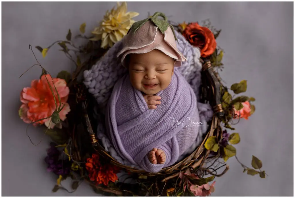 A newborn baby wrapped in a purple blanket, wearing a flower hat, smiles while lying in a nest surrounded by colorful flowers—a heartwarming scene perfect for Mommy And Me Classes In Milford CT.