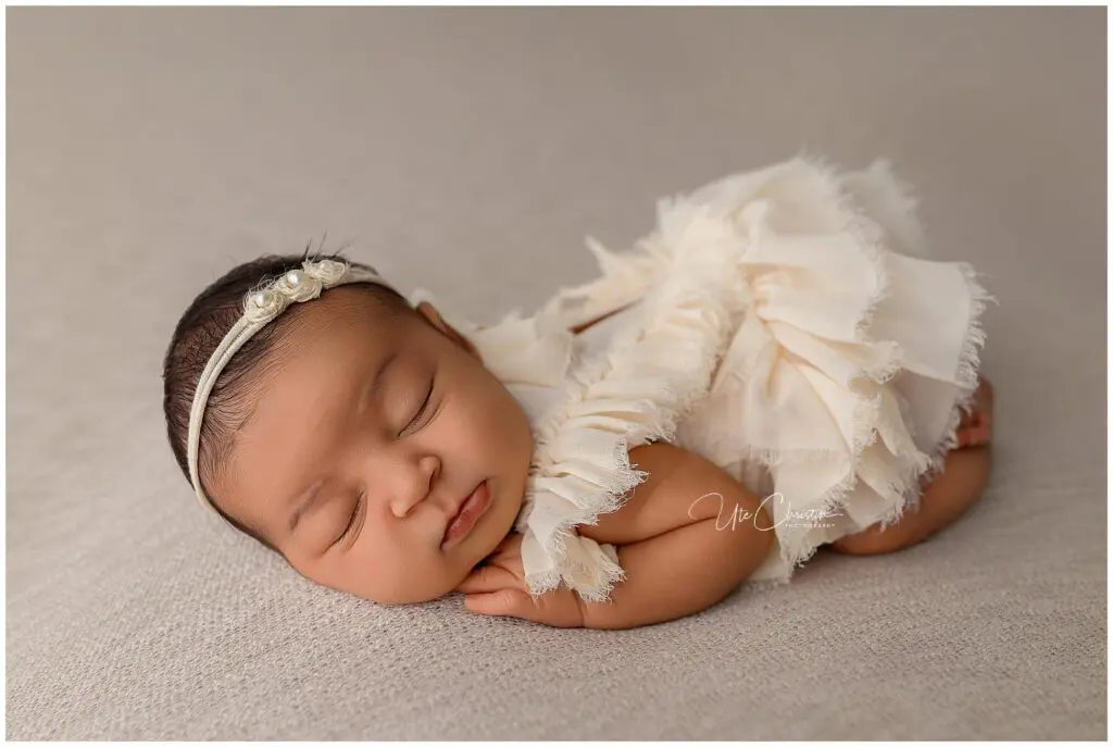 A newborn baby wearing a cream-colored ruffled dress and pearl headband sleeps peacefully on a soft beige surface with hands tucked under their cheek—perfect for precious moments before joining Mommy And Me Classes In Milford CT.